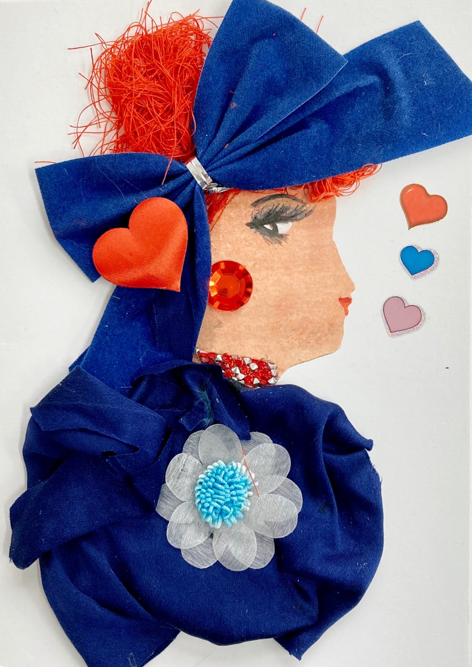 This card has. been given the name Holly. Holly wears a dark blue dress made of a lightweight fabric which has a large white flower pendant in the middle. She wears a large blue bow in her vibrant red hair, and on top of the bow there is a large red heart. Her earring is a large red gem, and her necklace is a cluster of small white and red diamonds. To the right of her, there are red, blue, and pink hearts.