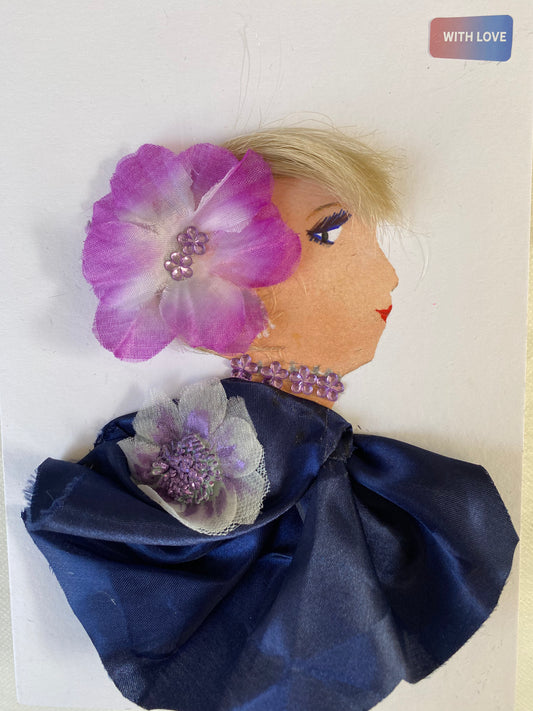 I designed this card of a woman named Dalston Doodle. She has a white skin tone and is wearing a stunning purple flower in her hair. She wears a silky blue blouse with a purple and white flower. She wears shiny purple jewellery.