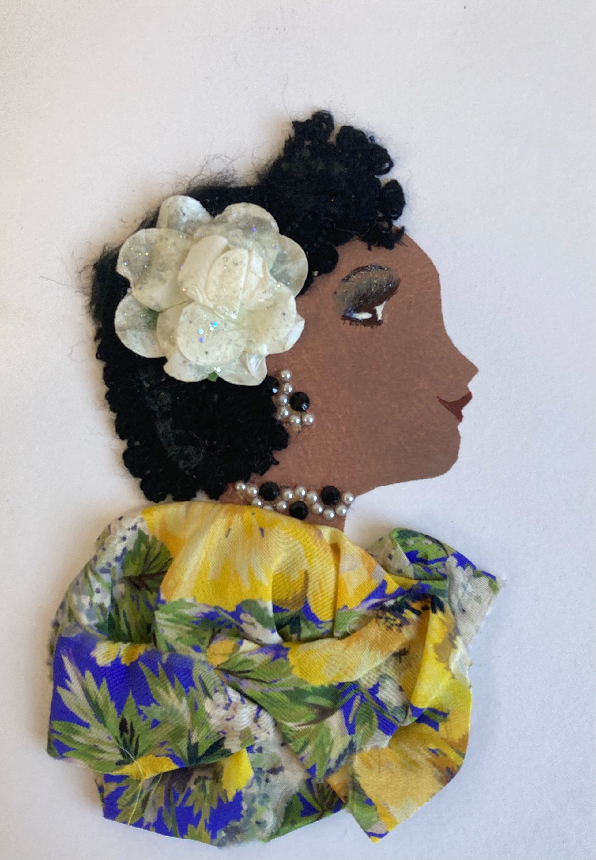This card is called Hattie. Hattie wears a floral print blouse with blue, yellow and green, and a white flower in her curly black hair.