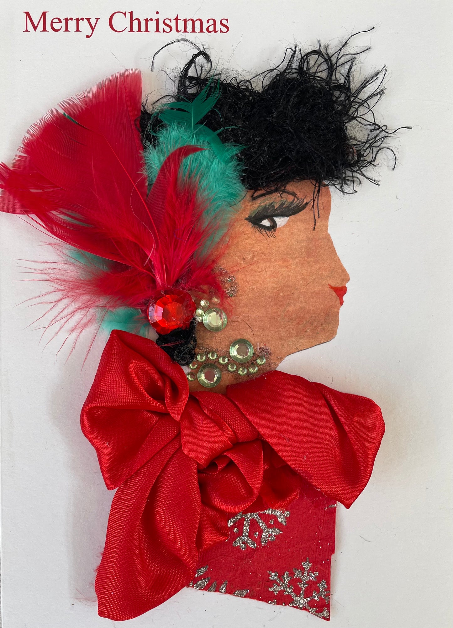 Handmade Christmas card featuring the side profile of a woman with curly black textured hair, decorated with red and green feathers and a red gem. She has red lips and green eye makeup. Her earring and neck accessory are made from green rhinestones. Her outfit is made of red fabric with silver snowflake patterns and is adorned with a large red satin bow. The background is plain white with “Merry Christmas” printed in red at the top left.
