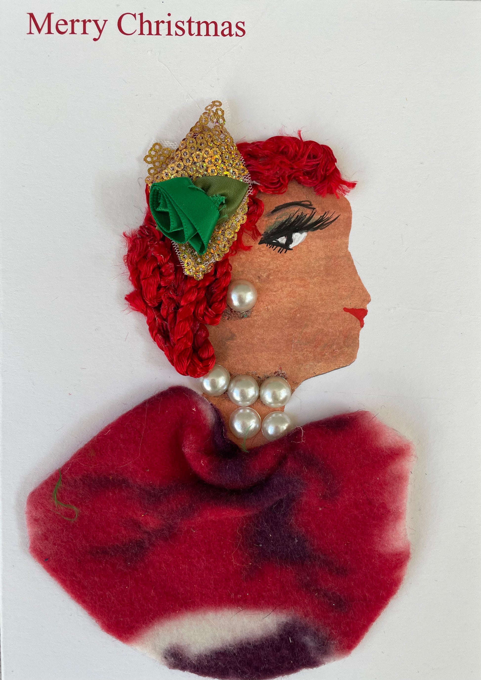 Handmade Christmas card featuring the side profile of a woman with bright red textured hair, decorated with a gold mesh accessory and a small green ribbon rose. She wears pearl embellishments on her ear and neck. Her outfit is made from maroon and white tie-dyed fabric. The background is plain white with the words "Merry Christmas" printed in red at the top left.