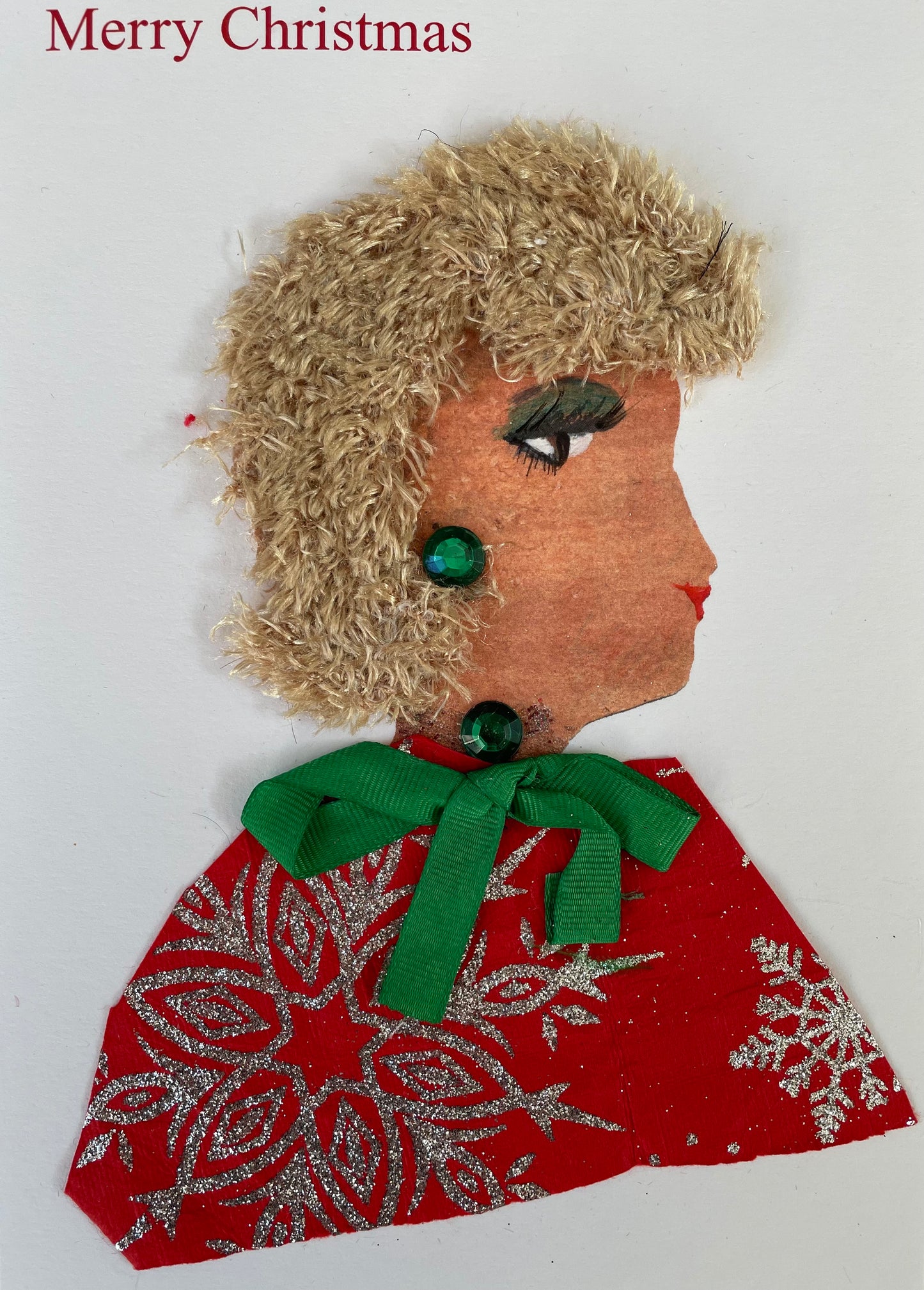 Handmade Christmas card featuring the side profile of a woman with curly, textured blonde hair, green eye makeup, and red lips. She wears green gem-like embellishments on her ear and neck. Her outfit is made from red paper with silver glitter snowflake patterns and is tied with a green ribbon bow. The background is plain white with the words "Merry Christmas" printed in red at the top left.