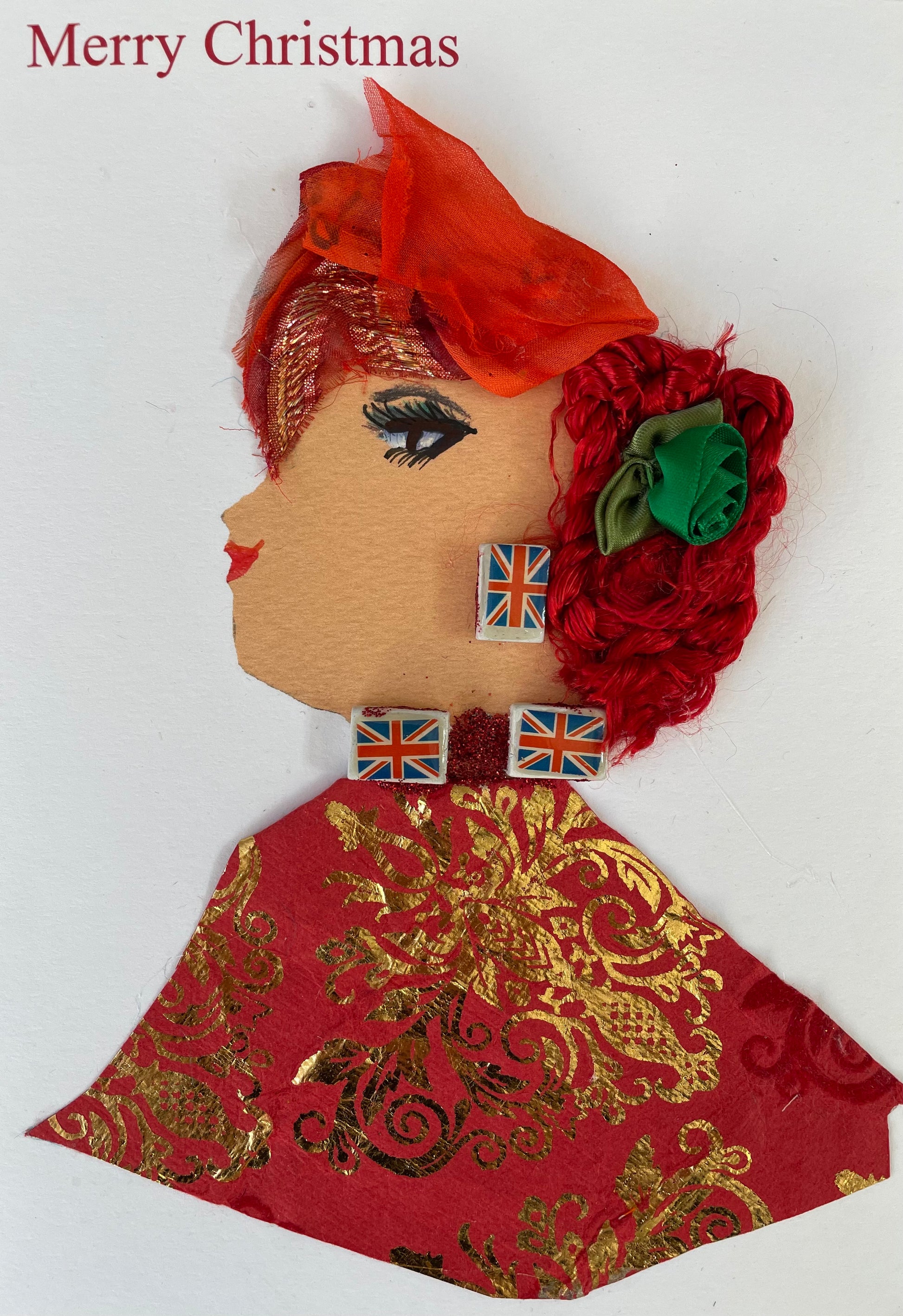 Handmade Christmas card featuring the side profile of a woman with painted features and red lips. Her hair is red and textured, adorned with a green ribbon rose and an orange fabric headpiece. She wears decorative blocks featuring the Union Jack as earrings and along her neckline. Her outfit is made from red paper with gold foil floral patterns. The background is plain white with “Merry Christmas” printed in red at the top left.