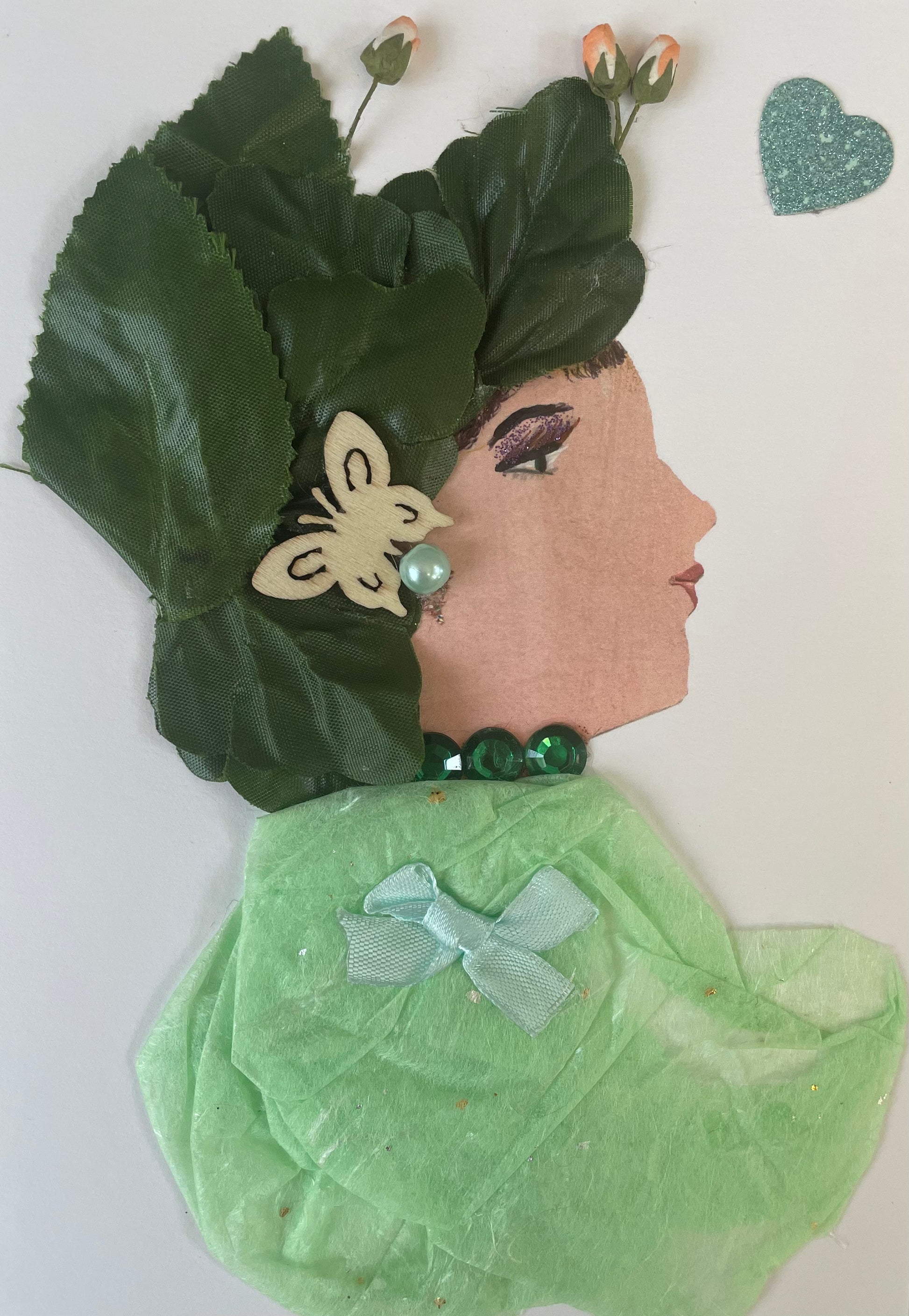 This handmade card called Celery Cyprus is wearing a light green blouse that she pairs it with a light blue bow and dark green necklace. She has a dark green leaf like headpiece with tulips coming out from the top and a butterfly bow.. There is a sparkly blue heart to the right of her.