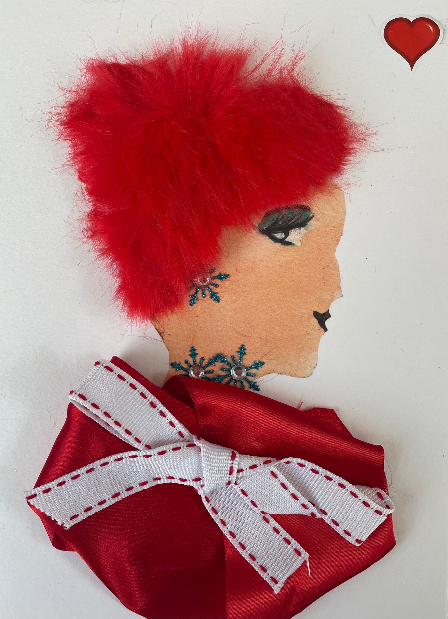 I designed this card of a woman called Victoria Valentine. She is a white woman who is wearing a red blouse with a white ribboned bow with red stitching at the edges. She has red vibrant hair. She has a matching blue diamanté snowflake designed necklace and earrings. There is a red heart in the right corner.