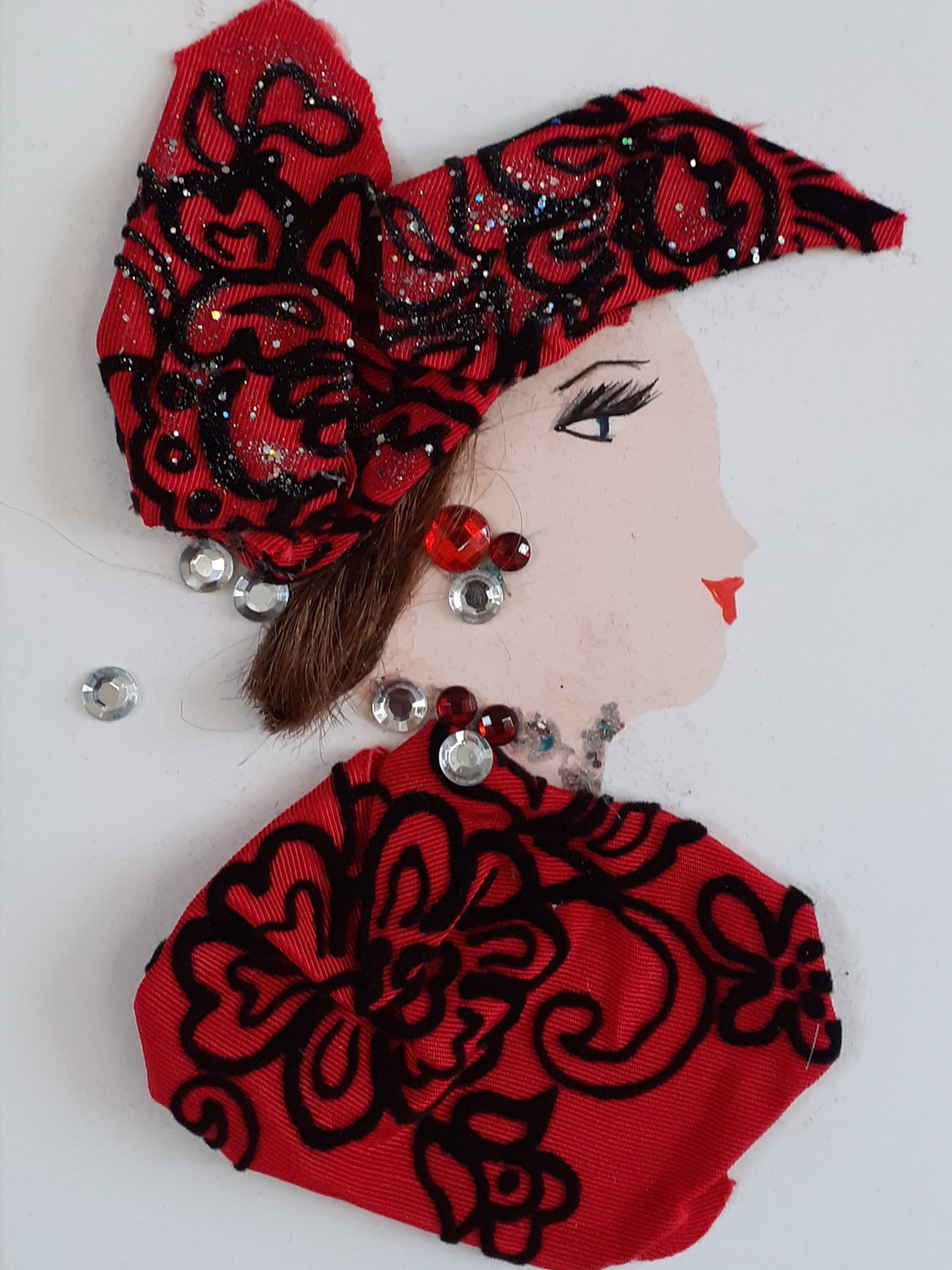 This card shows a woman wearing a red blouse and matching headdress which have black embroidery on them. Her short brown hair peeks out of the bottom of the headdress. She wears clear and red gem jewellery.