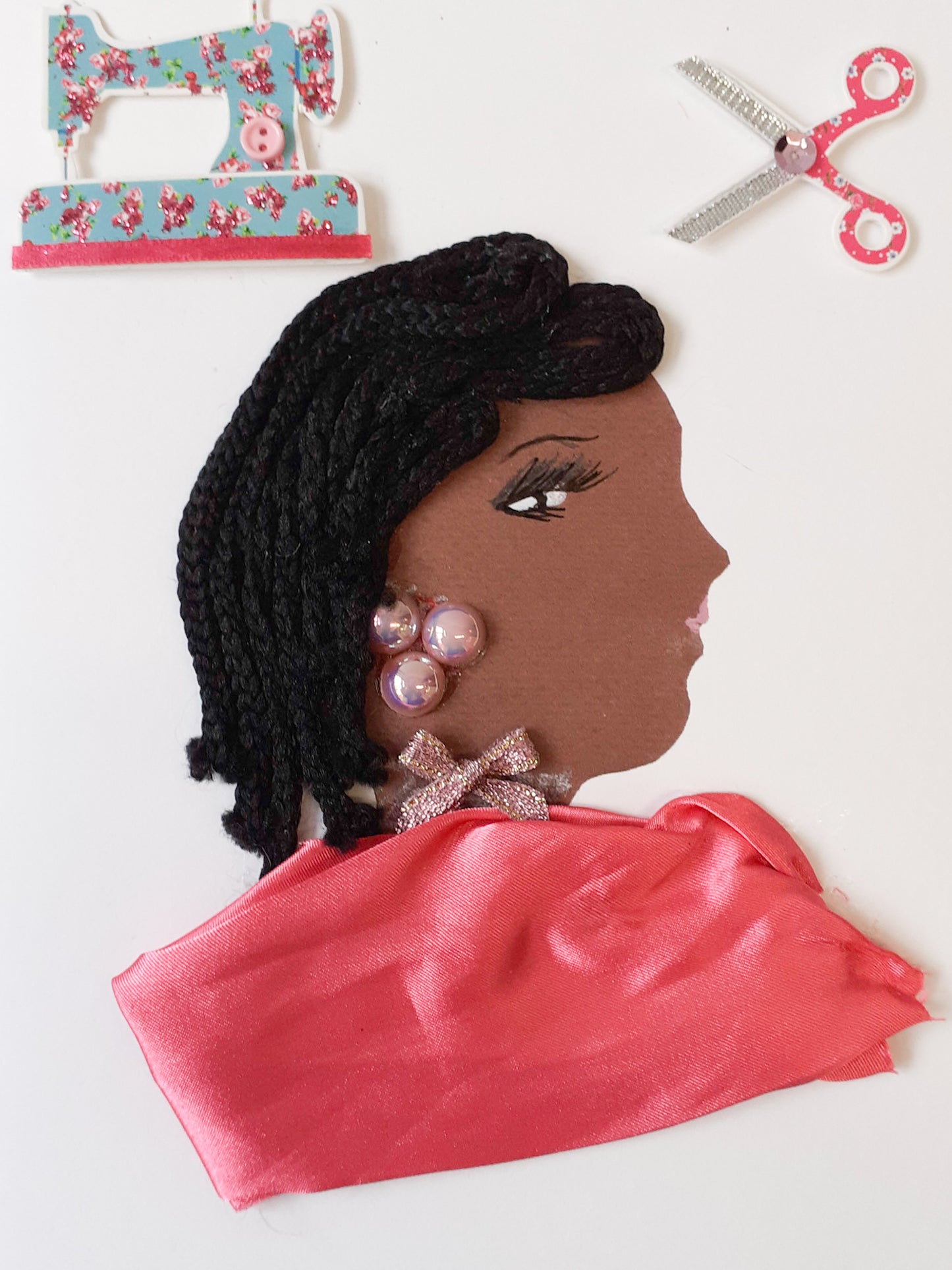 This card has been given the name Suzi Seamstress. Suzy wears a blush pink silk blouse and a small glittery bow around her neck. Her earrings are made of three pink iridescent pearls, and peek out of her long, black, braided hair. In the top left corner, there is a sticker of a sewing machine which has a floral pattern on it. In the top right corner, there is a sticker of a pair of stickers that have a jewel on them.