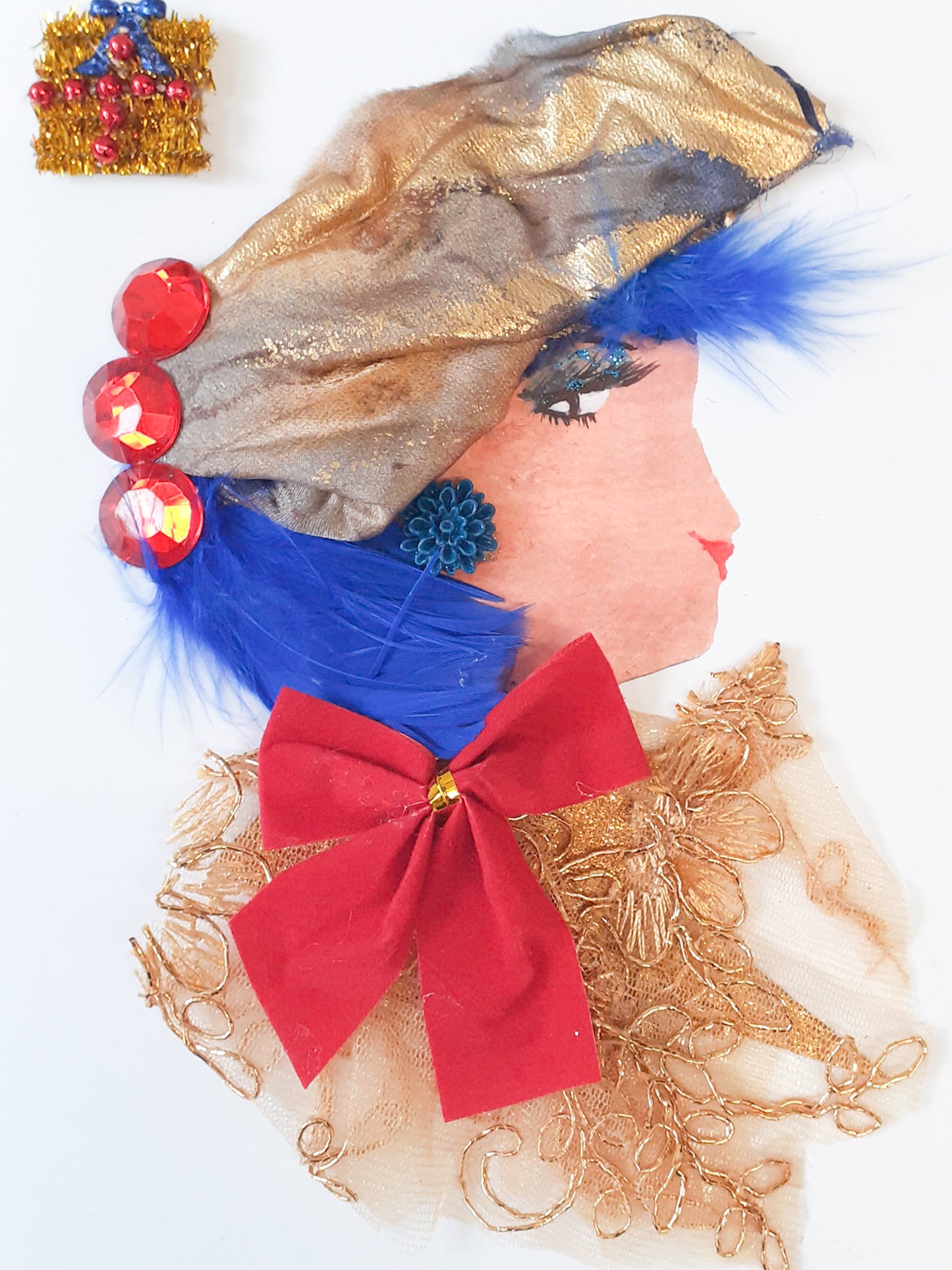 This card has been given the name Snowflake Court. She wears a gold fabric blouse with a giant red bow on it, she has blue feathery hair, and wears a gold headdress in it. In the top left corner, there is a sticker of a present.