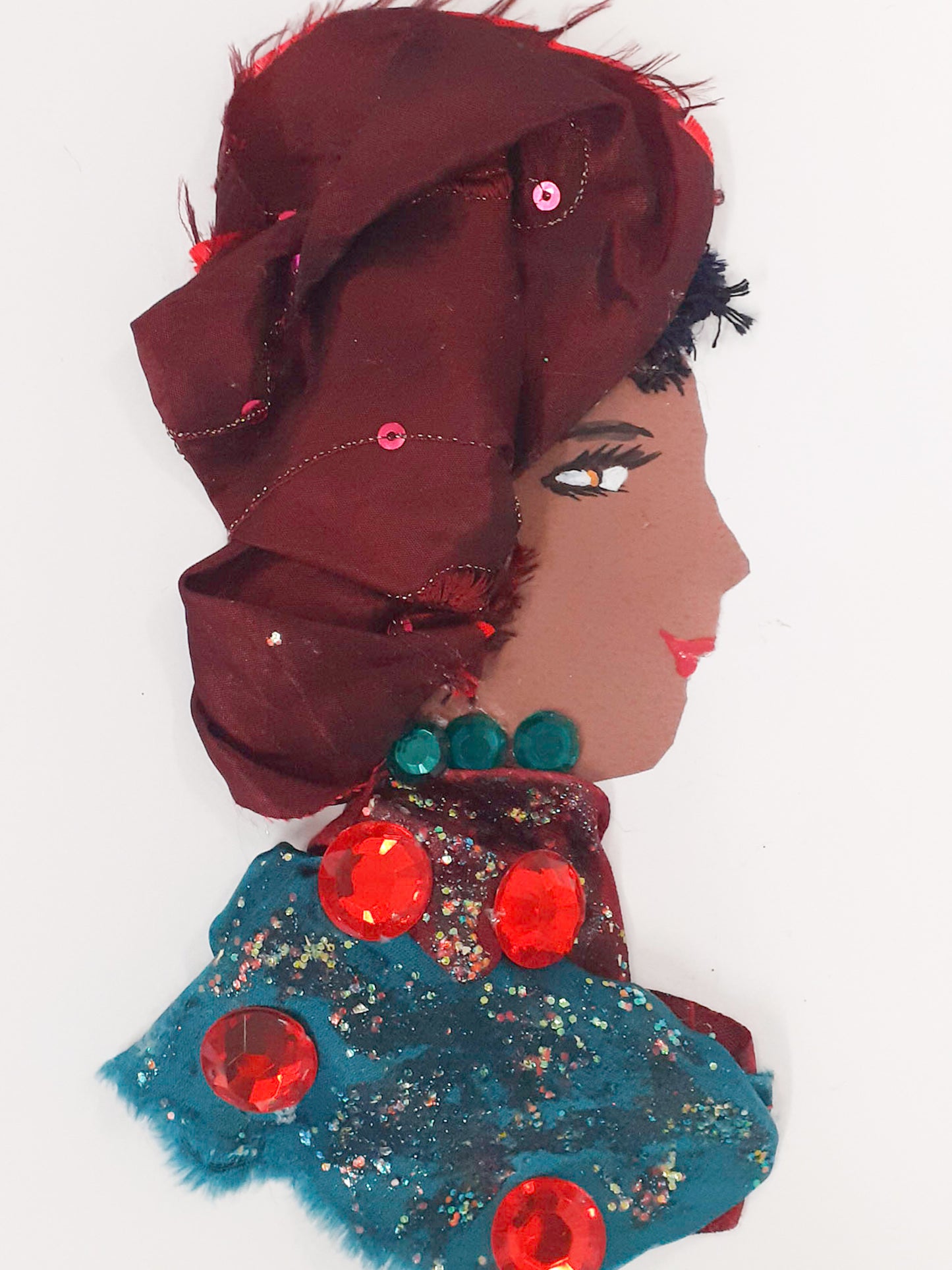 This card has been given the name Miss Gemstone. She wears a green blouse with glitter details and large red gemstones on her dress. She wears a dark red headdress with sequins on it.