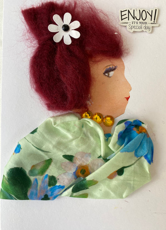 This is a handmade card of a woman named Merry May. Her deep burgundy hair is styled high and decorated with a white flower featuring a black centre. She wears a necklace made of bright yellow gems. Her outfit is made from soft green fabric with a floral print in white, blue, and hints of orange. Her face is drawn in profile with delicate features and red lips. In the top corner, a sticker reads, “Enjoy! It’s your special day.”
