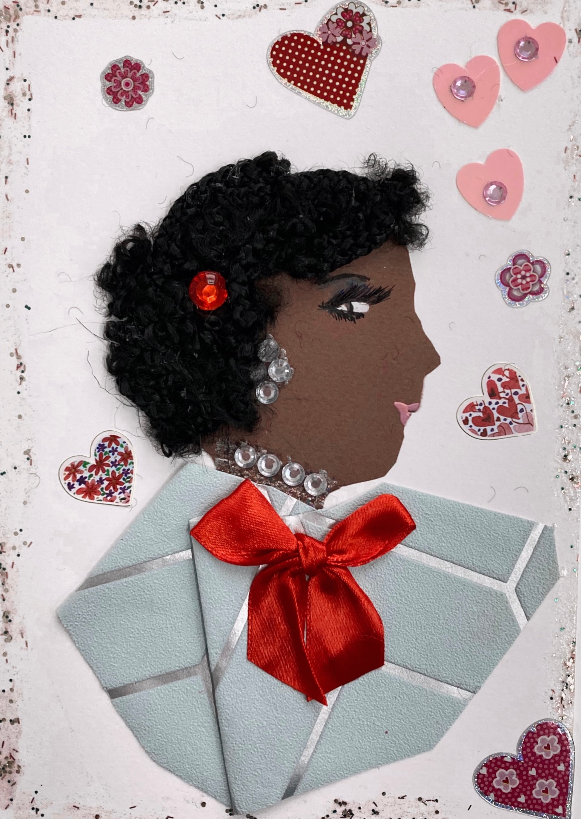 This is a handmade card of a woman named Miss Ruislip Gardens. Her hair is styled with tight black curls and accented with a small red gem. She wears silver gem earrings and a matching necklace. Her outfit is made from soft blue paper with a silver stripe pattern, finished with a bright red satin bow. The card is decorated with stickers of hearts and flowers in pink, red, and patterned designs.
