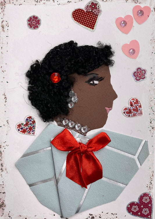 This is a handmade card of a woman named Miss Ruislip Gardens. Her hair is styled with tight black curls and accented with a small red gem. She wears silver gem earrings and a matching necklace. Her outfit is made from soft blue paper with a silver stripe pattern, finished with a bright red satin bow. The card is decorated with stickers of hearts and flowers in pink, red, and patterned designs.