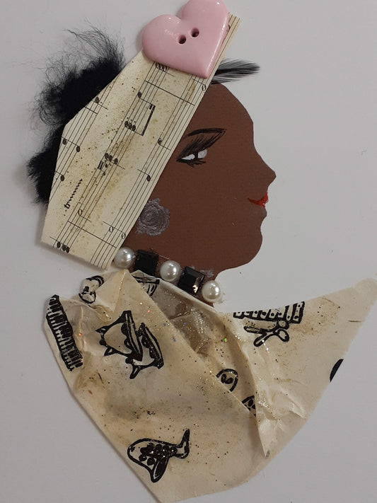 I handmade this card of a woman named Musical Mum. She’s shown in profile with a warm smile, soft lashes, and a printed silver earring. Her headwrap is made from aged sheet music and topped with a glossy pink heart-shaped button. She wears a beaded necklace of pearls and dark squares, and her outfit is crafted from beige fabric printed with black fish and musical motifs, lightly dusted with glitter.