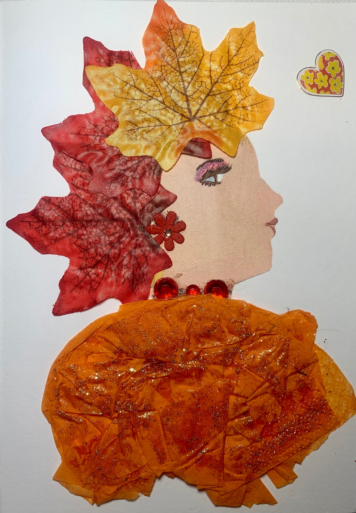 This card is of a woman called Betty. She wears a dress made of an orange paper material with gold sparkles on top. She wears a red necklace made of gemstones. On top of her head there are three oak leaves which have turned red, orange, and yellow. She wears a red flower earring. In the background, there is a small heart which has orange and yellow flowers on it.