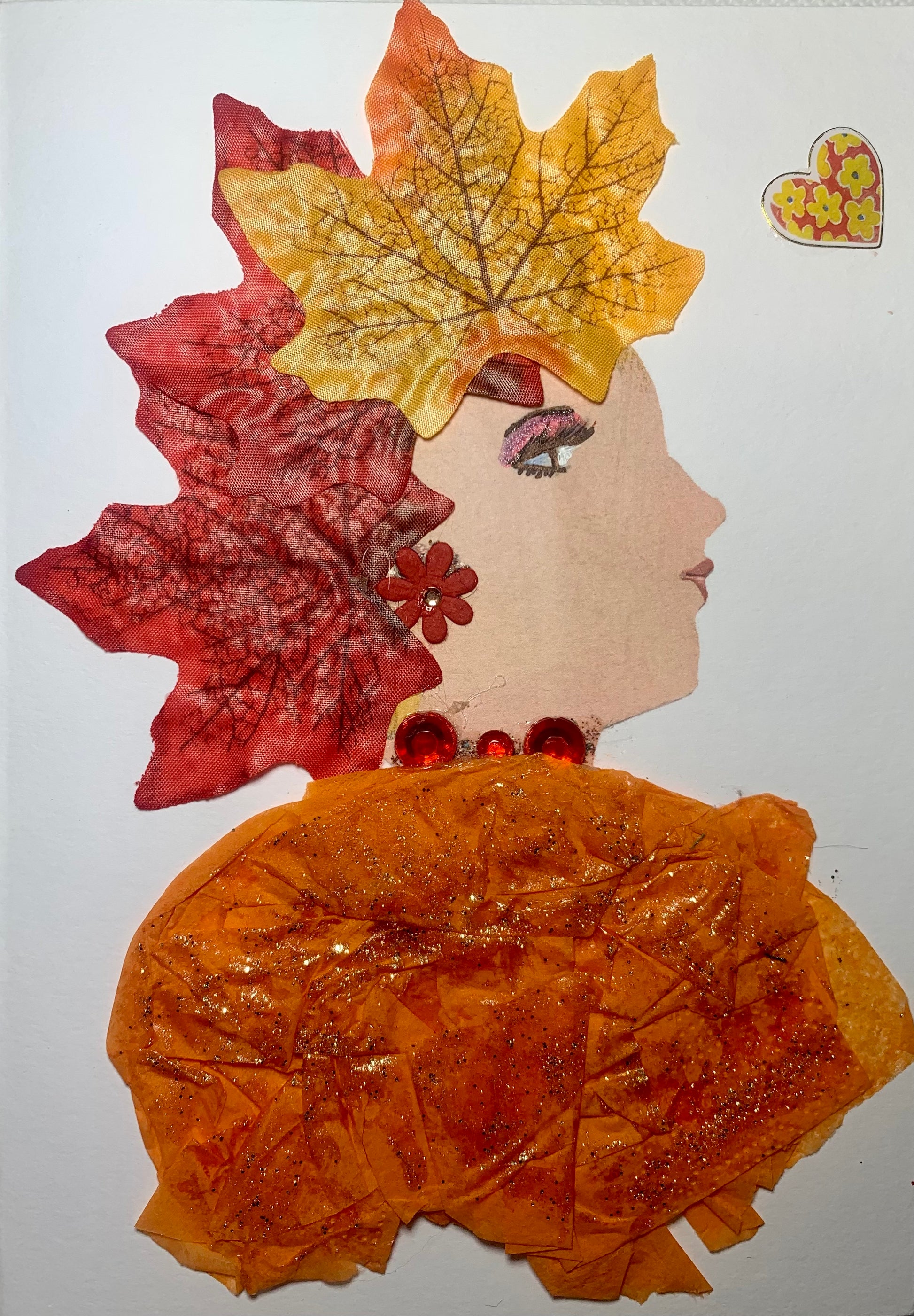 This card is of a woman called Betty. She wears a dress made of an orange paper material with gold sparkles on top. She wears a red necklace made of gemstones. On top of her head there are three oak leaves which have turned red, orange, and yellow. She wears a red flower earring. In the background, there is a small heart which has orange and yellow flowers on it.