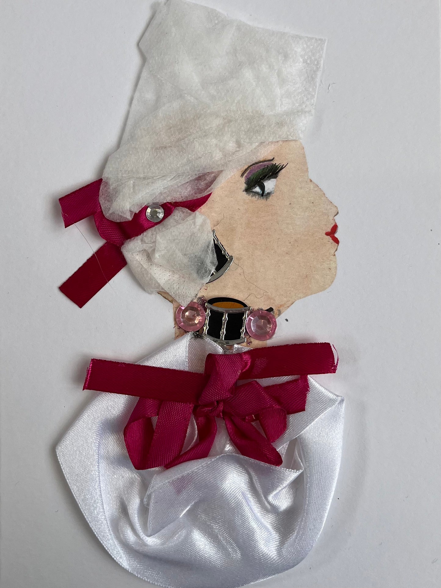 This card has been given the name Tessa. Tessa wears a white silky blouse with a magenta bow on the front. Her necklace is made up of a small black chain and two pink gems. She wears a white headscarf with a matching magenta ribbon in it.