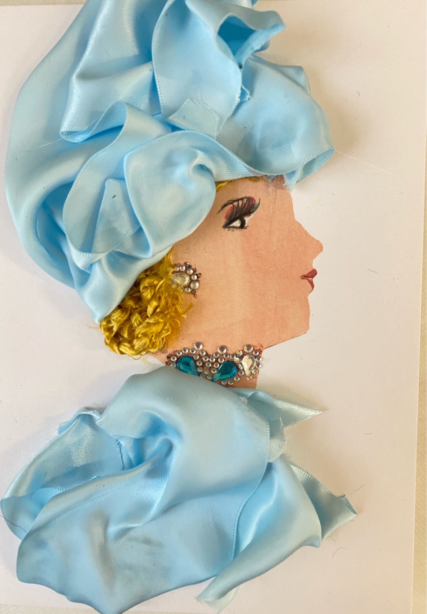 I designed this card of a woman named Emma Jivmark Blue. She has a white skin tone and is wearing a divine silk blue hat matched with an angelic silk blue blouse. Her outfit is paired with dazzling turquoise and silver jewellery.