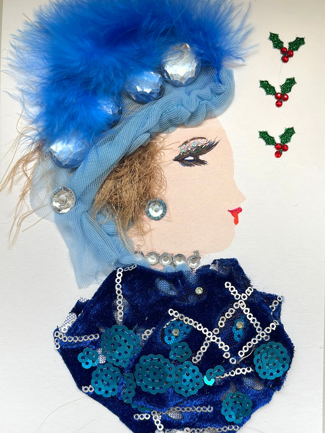 This card has been given the name Adaya. Adaya wears a dark blue blouse with ocean blue sequin circles on it, and lines of silver sequin. She wears a diamonte gem necklace, and a blue gem earring. In her light blonde hair she wears a blue feathery headdress with large blue gems in it. In the righthand corner, there is mistletoe. A part of what I sell is not just my cards but the experience of choosing a card as well so visit the website for more options.