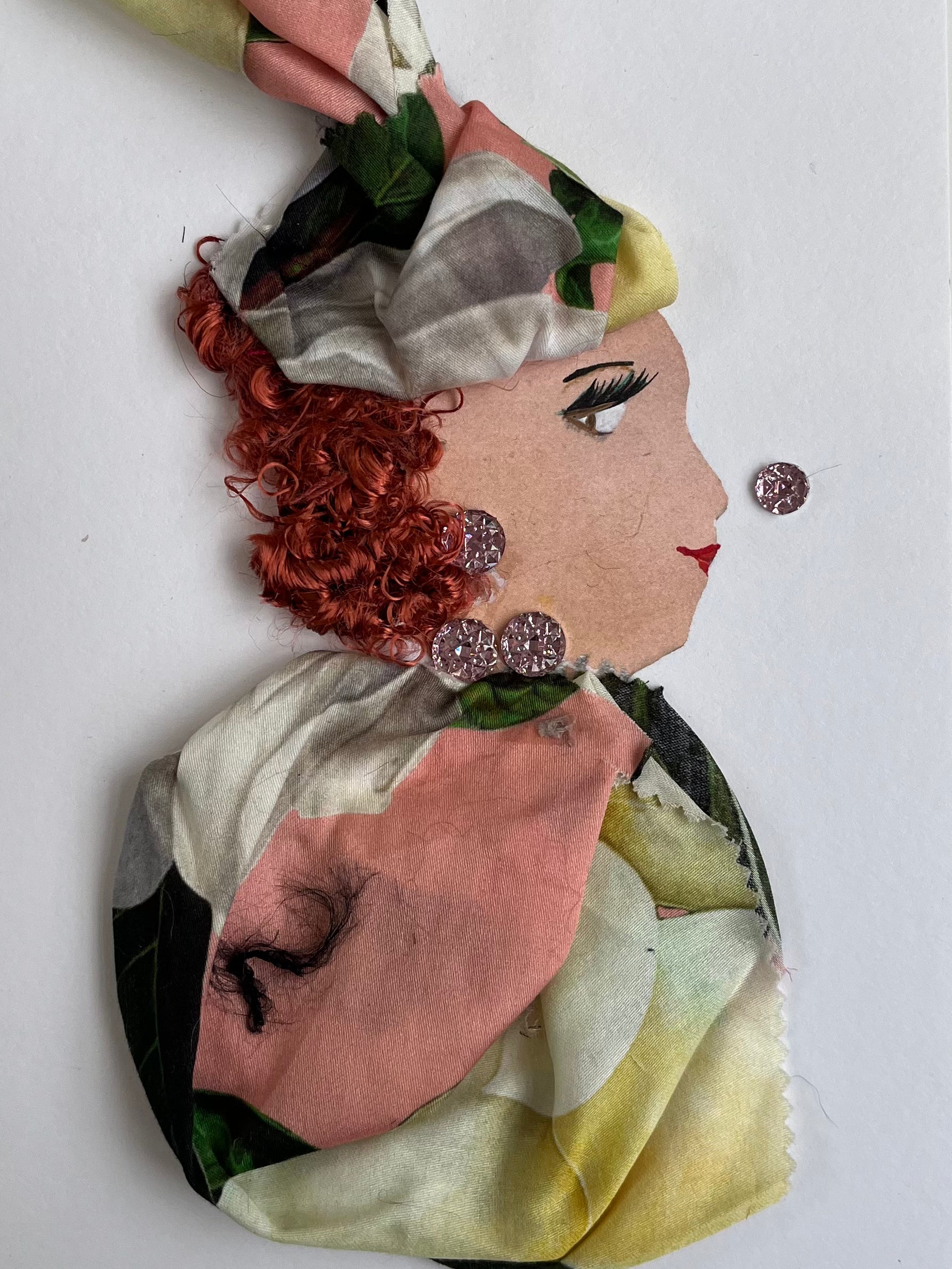 Kendall wears a floral printed headdress and blouse, both which are a pink and light yellow pattern. Her red hair is short and curly, and she wears pink gemstone jewellery.