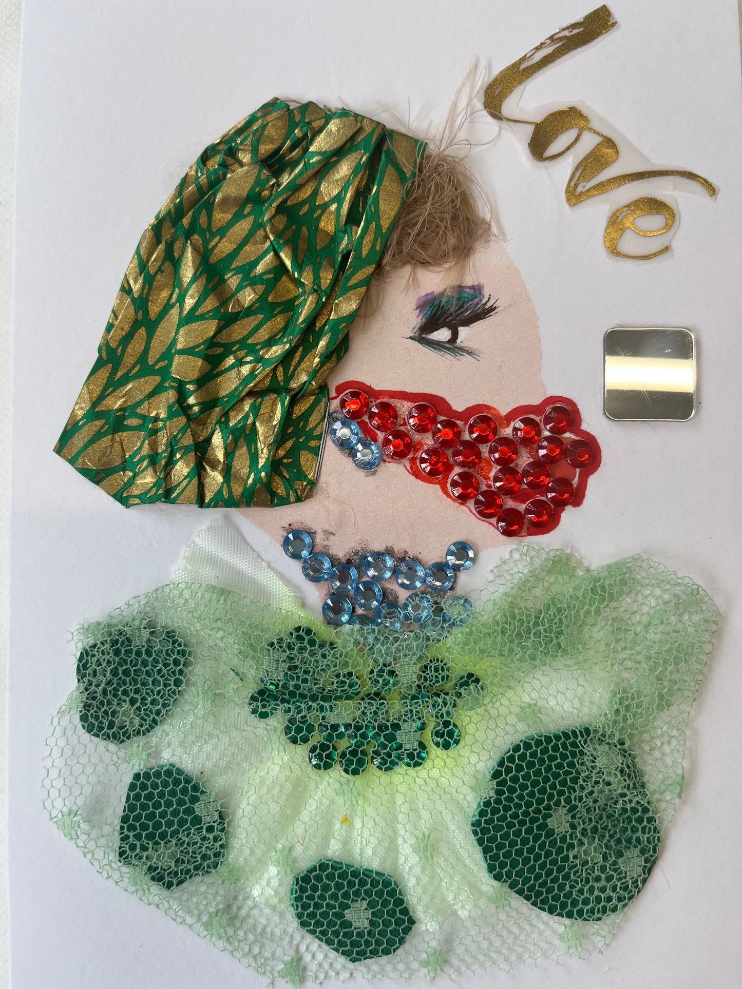 This card has been given the name Love Mask. Love wears a green blouse with small forest green ovals underneath a lighter green sheer mesh. Her necklace and earrings are both made of light blue gems. She wears a red gem mask over her mouth. In her light brown hair, she wears a green headscarf with a gold leaf pattern. In the top right corner, it sayd "love" in gold writing. Underneath, there is a small square mirror.