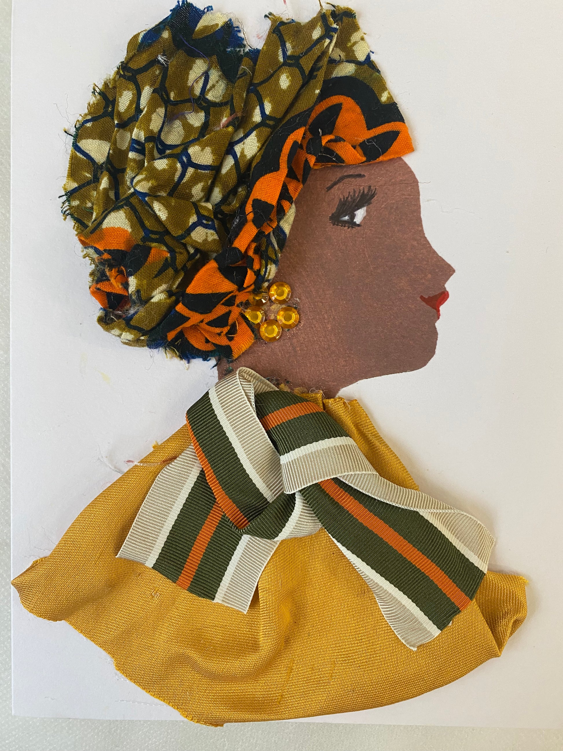I designed this handmade card of a woman named Golden Gabriella. She has a brown skin tone wears an orange and green ankara hat. She wears an orange and green stripe bow with a graceful golden blouse. She wears beautiful gold earrings.