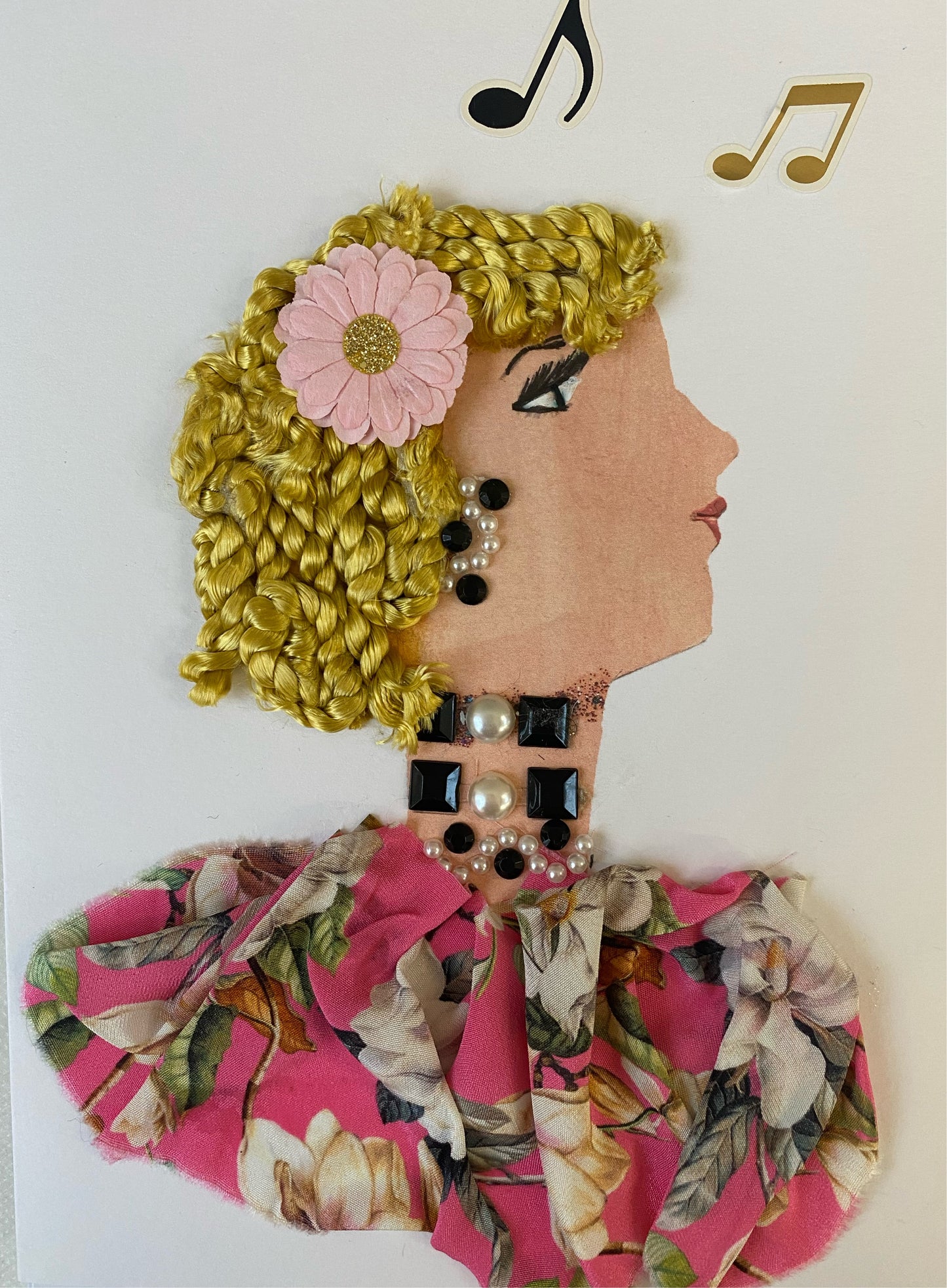 This is a handmade card of a woman named Nancy Blushes. She has a white skin tone and she wears a pretty pink flower in her hair. She wears a pink floral blouse with black and pearl jewellery. In the corner there are music notes.