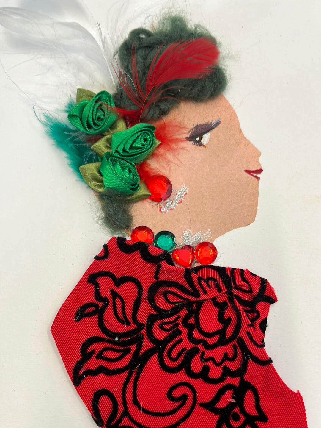 I designed this card of a woman who has a white skin tone. She is wearing a red blouse with black floral embroidery. Her hair is green yarn, and has lots of green flowers in it. She wears a red and white feather in her hair as well. She wears red, green, and silver jewellery.