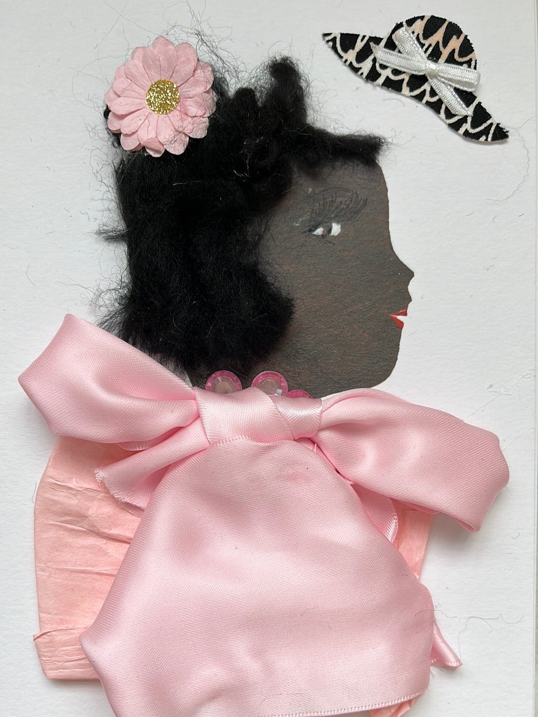 This card is of a woman given the name Hattie. Hattie wears a baby pink blouse that is made of a paper material, and there is a large silky pink bow on the neck. She wears a pink gem necklace, and a pink flower with a gold shimmery center in her short black hair. On the top right corner, there is a black and white floppy hat sticker which has a small white bow on it. A beautiful card  to frame for your home or the perfect gift for someone to celebrate