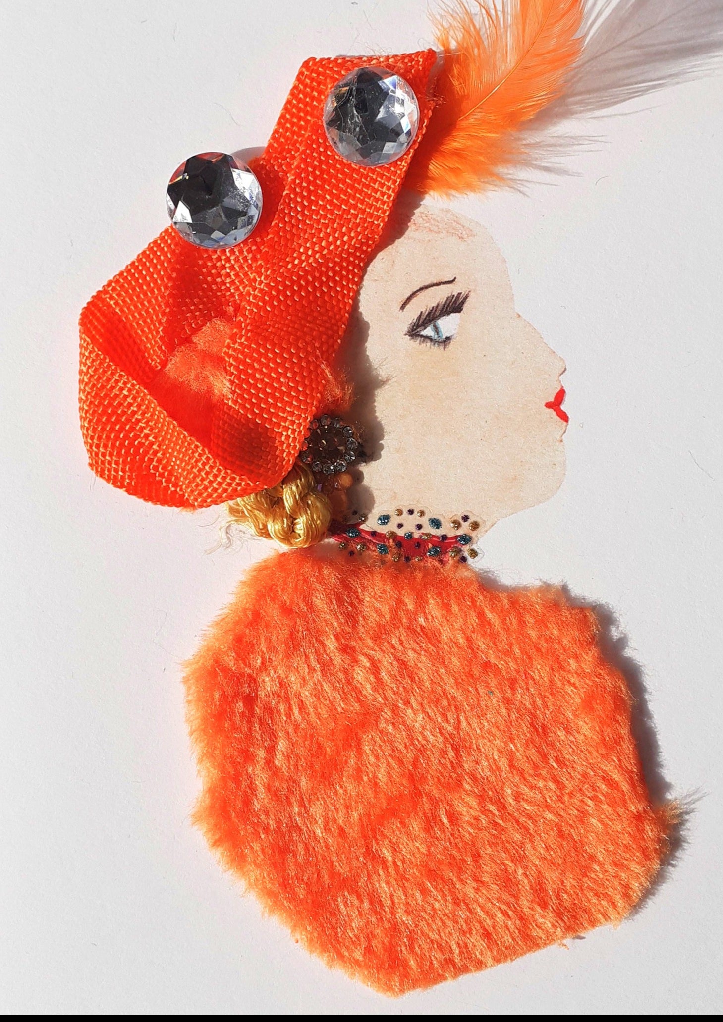 This card is called Olivia. She wears an orange fur blouse and a headress that matches in colour. There are two large crystal gems in her blonde hair as well as a feather.