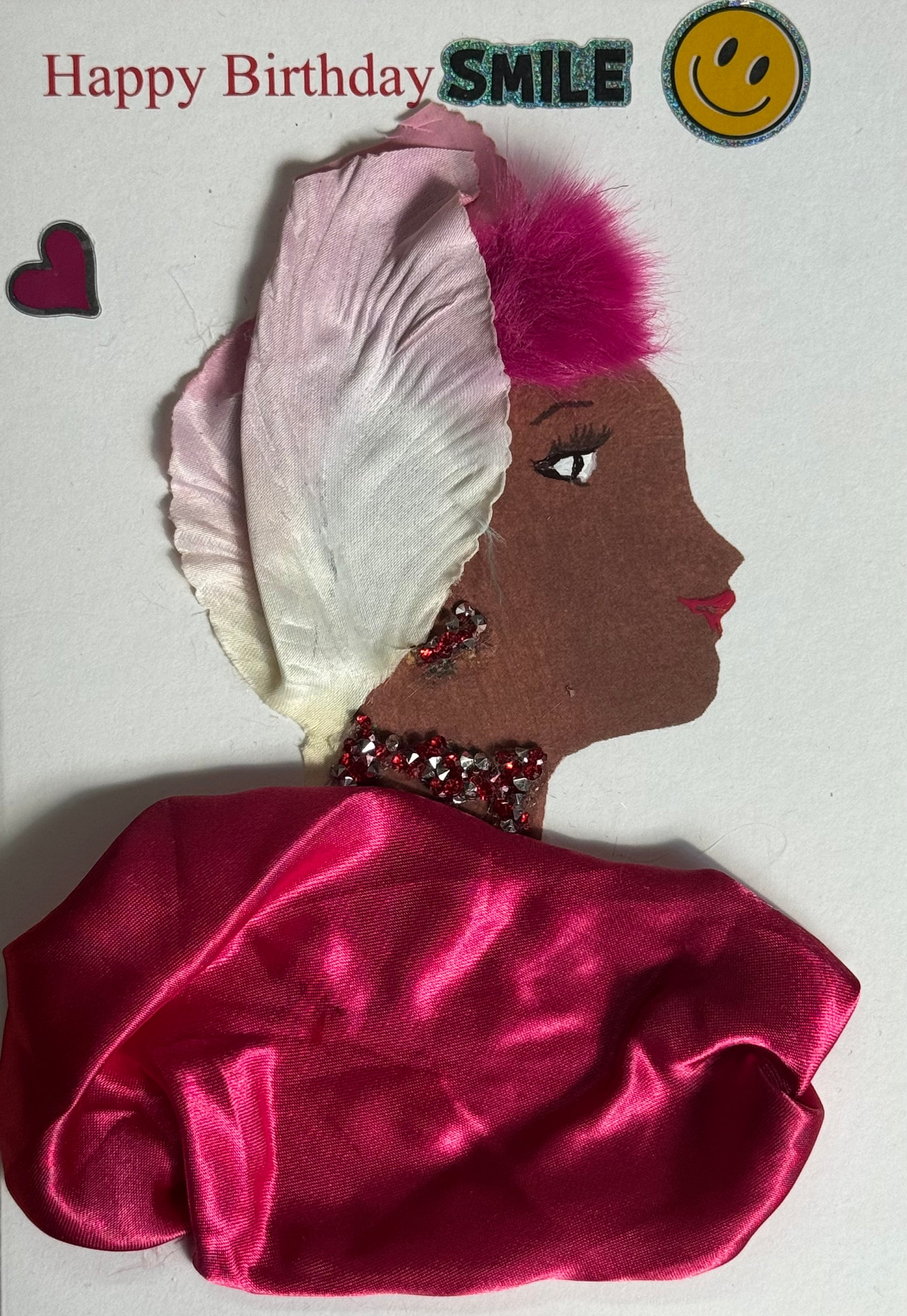 A handmade birthday card showing a woman with dark brown skin in side profile. She wears a white petal-shaped headpiece with a bright pink fluffy accent. Her jewelry includes a red gem necklace. Her wrap is made from shiny hot pink satin fabric draped across her shoulders. Above her head are a “SMILE” sticker, a yellow smiley face, and a small pink heart. “Happy Birthday” is printed in red on a white background.