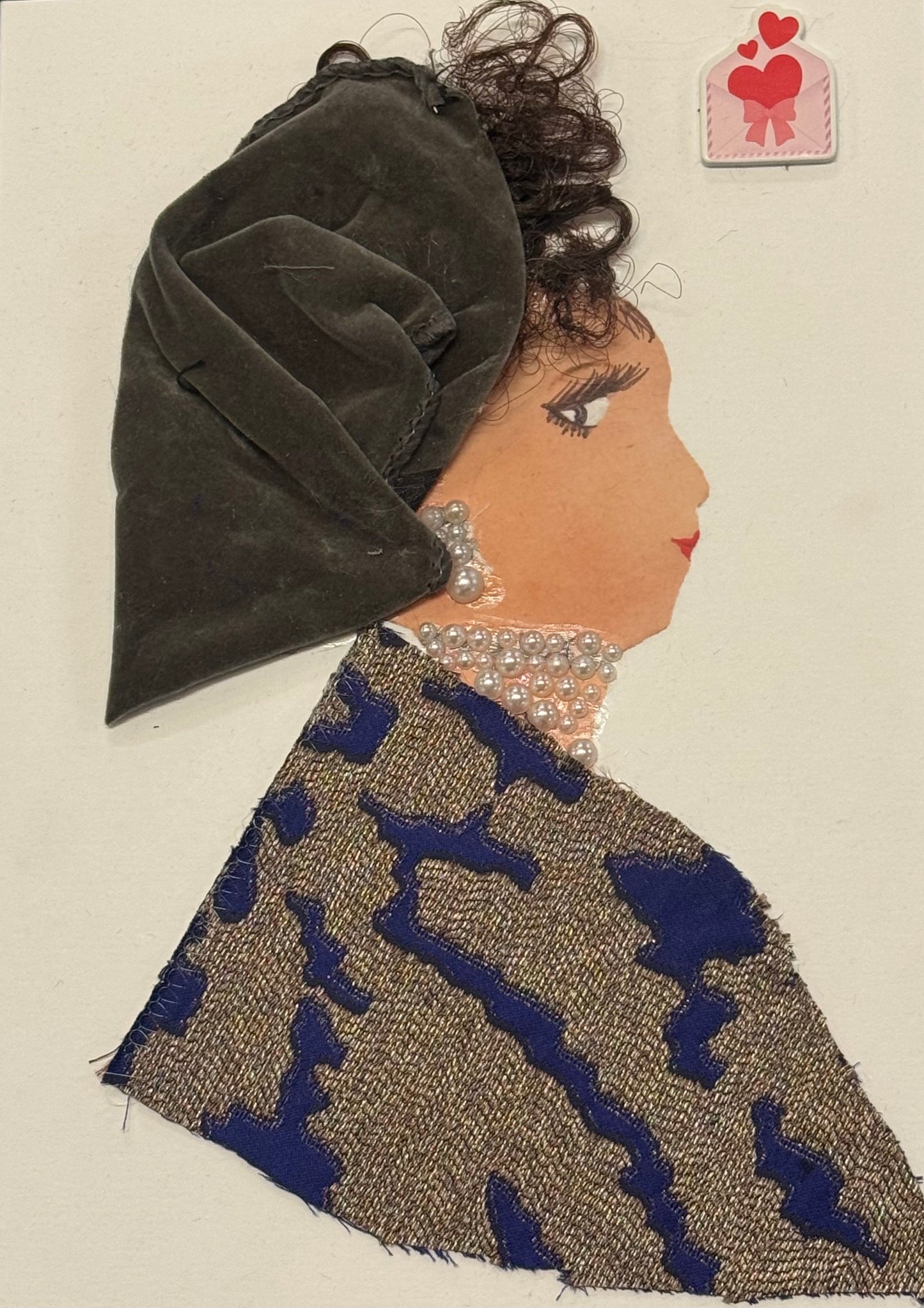 A side-profile collage of a woman with a warm medium skin tone against a plain white background. She wears a large, draped velvet headwrap in a deep grey color, with visible folds and a soft, matte texture. Dark curly hair emerges slightly from beneath the wrap. Her outfit consists of a textured fabric in beige tones with bold, abstract blue patterns. Around her neck and ear are layered pearl embellishments forming jewelry. Her facial features are drawn with a defined eye, lashes, and a subtle lip. 