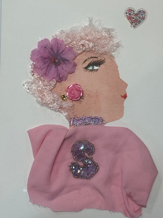 Handmade greeting card that can double as a piece of art, featuring a side-profile illustration of a woman with textured light pink curls adorned with a purple flower. She wears a pink fabric garment with a glittering jeweled “S” brooch, a sparkly choker, and pink rose earrings, all set against a clean white background.