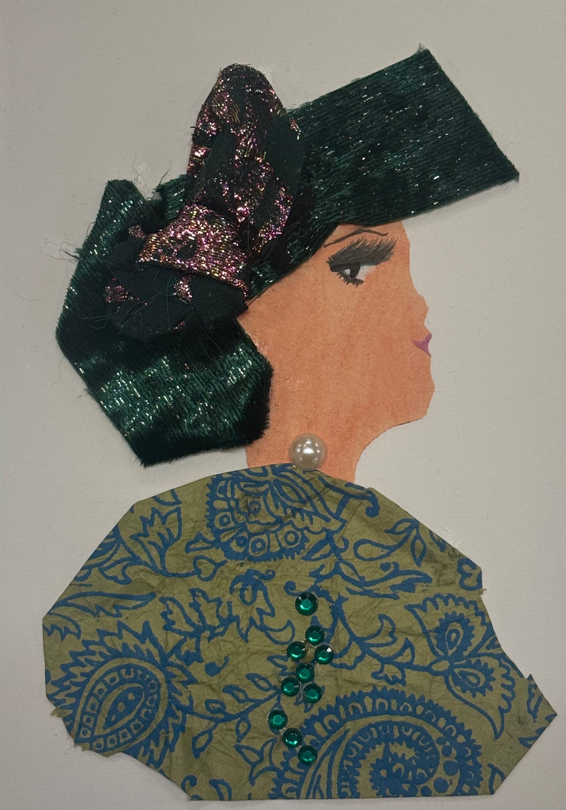 Handmade greeting card that can double as a piece of art, featuring a woman in profile wearing a large emerald green bow headpiece. She wears a patterned green, bold, and gold outfit with small green gems and a pearl detail. The design uses textured fabrics and decorative embellishments.