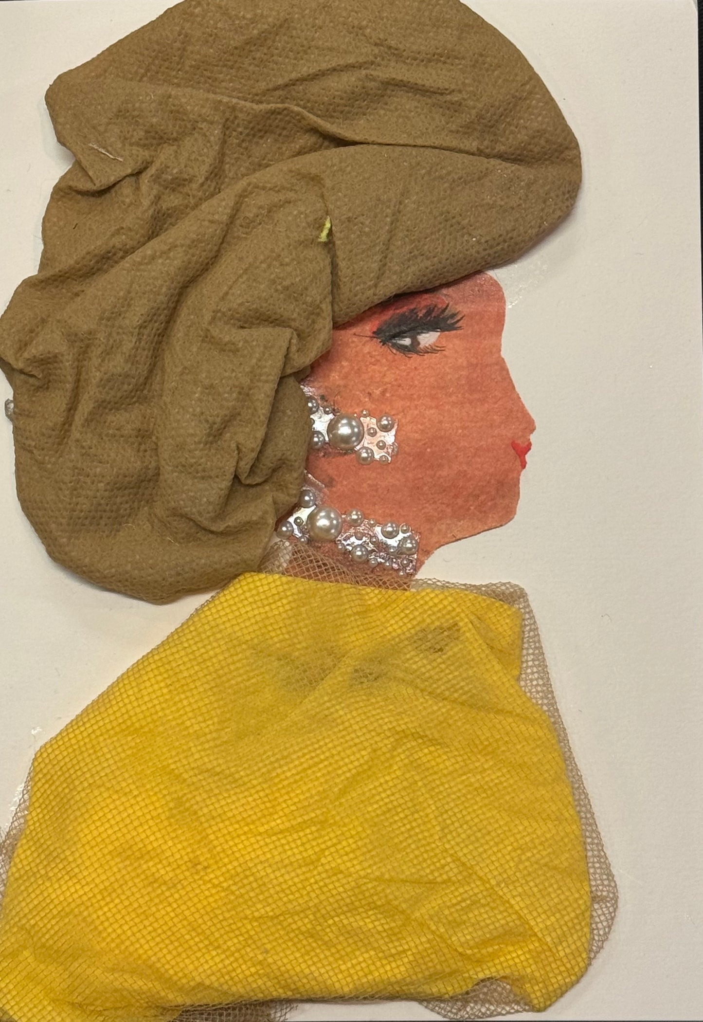 Handmade greeting card that can double as a piece of art featuring a side-profile silhouette of a woman with a warm orange-brown complexion. She wears a textured tan headwrap made from soft fabric and a draped yellow shawl across her shoulders. Her jewelry consists of pearl-like embellishments forming earrings and a necklace. Her facial features include defined lashes and soft red lips, all placed against a plain white background with layered fabric textures creating dimension.