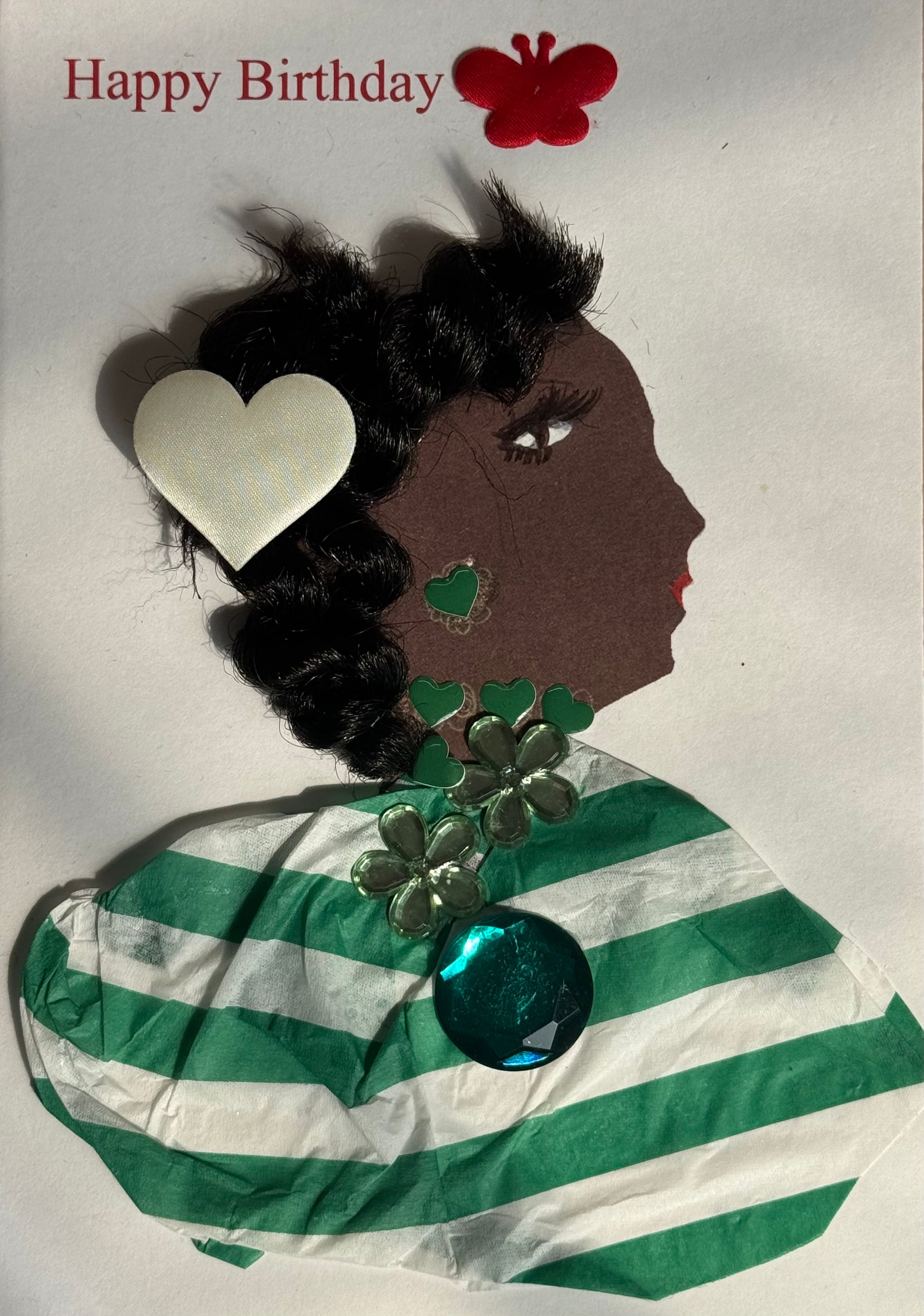 A handmade birthday card showing a woman with dark brown skin in right-facing profile. Her hair is styled in sculpted black curls with a large white heart-shaped hair clip on the side. She wears green heart-shaped earrings and a layered necklace made of green gem flowers with a large teal circular jewel hanging at the bottom. Her outfit is created from green and white striped textured material wrapped around her shoulders. A red butterfly sticker appears above her head, and “Happy Birthday” is printed in re