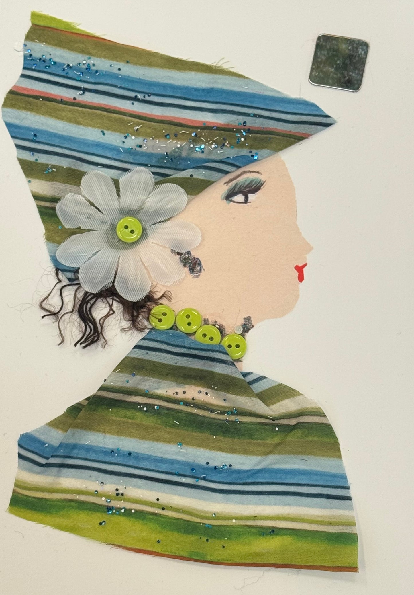 Handmade collage greeting card showing a right-facing side profile of a light-skinned woman wearing a headwrap and outfit made from striped fabric in shades of green, blue, and hints of white. The fabric includes small glitter accents scattered across it. A large white fabric flower with a green button center is attached near her ear, serving as a prominent accessory. Her hair appears as short, curly black strands peeking out beneath the headwrap.