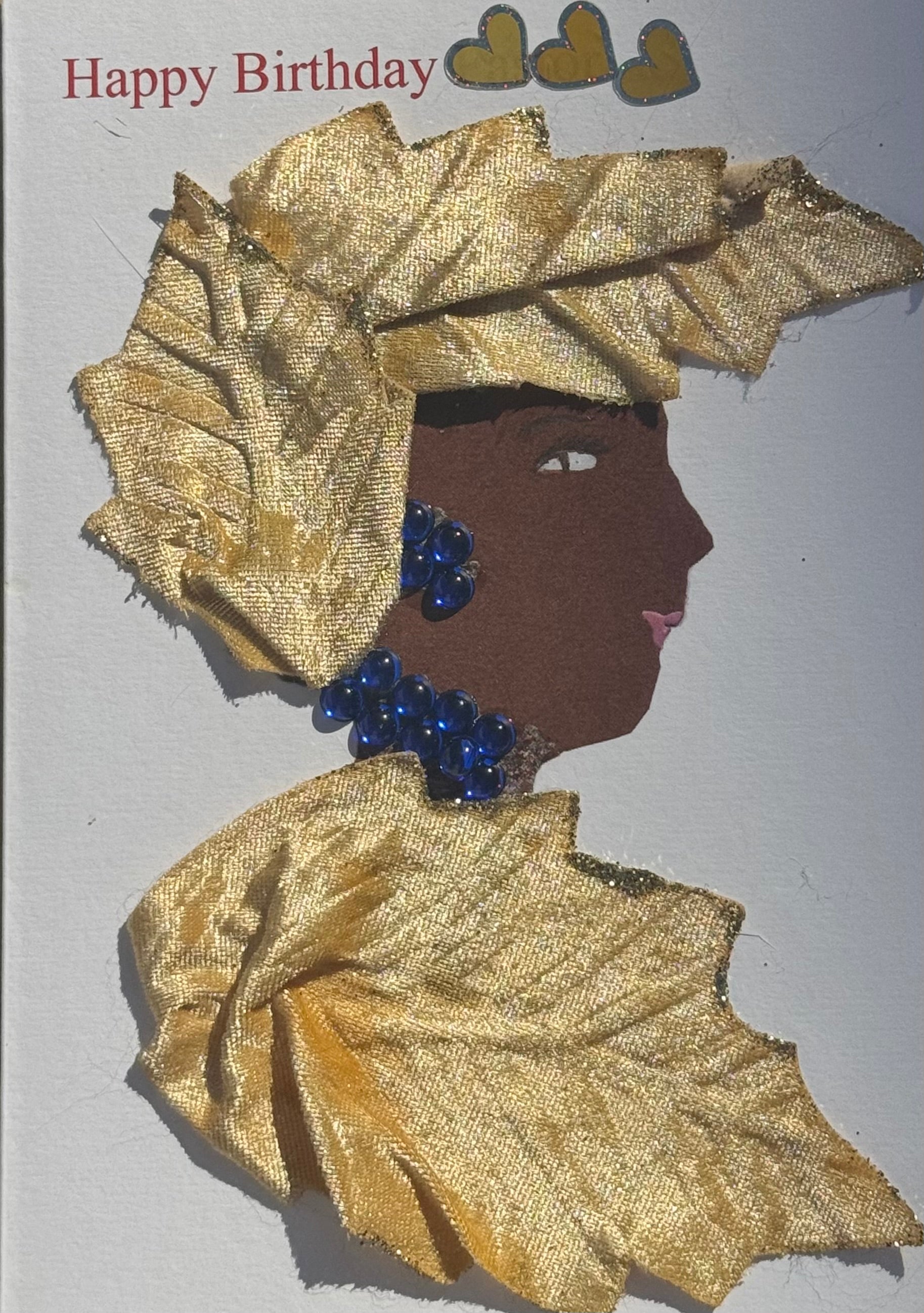 Handmade greeting card that can double as a piece of art featuring a side profile collage of a woman wearing a textured gold headwrap and matching gold garment, blue beaded necklace and earrings, soft pink lips, and decorative hearts above with “Happy Birthday” text on a white background.