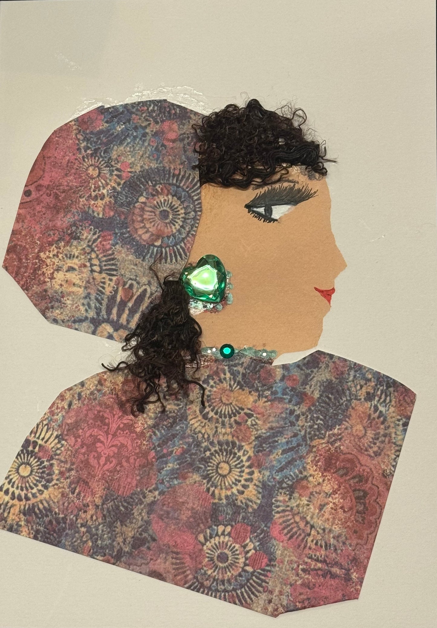 Handmade greeting card that can double as a piece of art featuring a side-profile portrait of a woman with dark curly textured hair partially tucked under a patterned headscarf made from fabric with intricate circular and floral designs in muted red, blue, and gold tones. She wears a matching garment and is adorned with a large emerald-green gem earring and coordinating necklace accents. She is defined lashes and red lips, and the overall composition combines fabric, paper, and embellishments. 