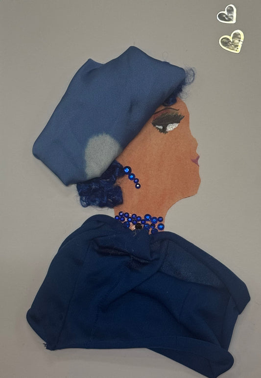 Handmade card that can double as a piece of art showing a woman wearing a deep blue bonnet with blue curly hair underneath. With this, she wears matching shiny blue beaded jewelry. She is also depicted wearing a dark blue shawl for her top. This design features cool tones as well as decorative embellishments like the two small silver hearts in the top left that give the card a calm and elegant feel. 