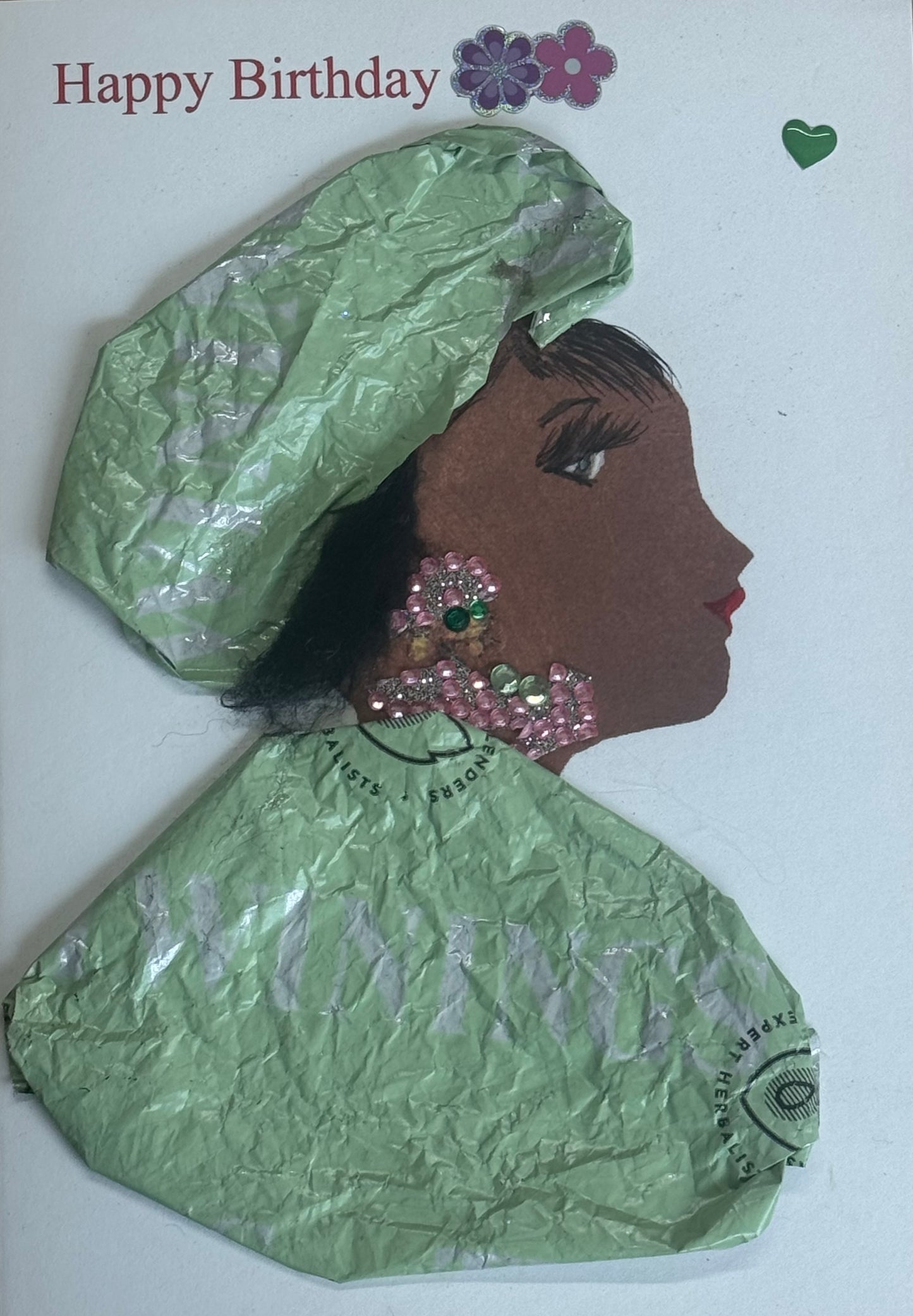 A handmade birthday card showing a side-profile illustration of a woman with warm brown skin wearing a large mint green textured headwrap made from crinkled tissue paper. She has pink gem earrings layered with a green central jewel and matching rhinestone necklace details. She wears a matching mint green shawl and headwrap made from a wrapper material.  The background is white with “Happy Birthday” printed above and small flower stickers near the top. 
