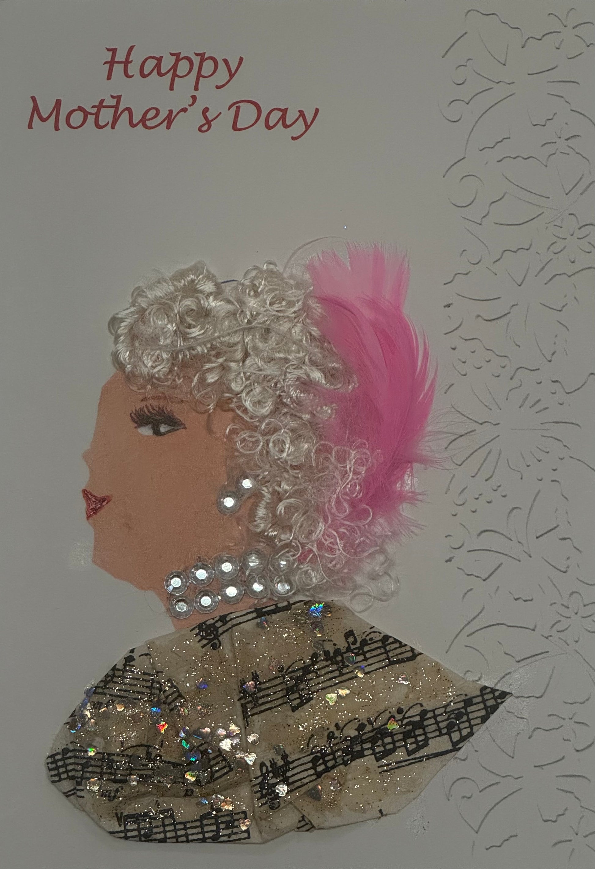 A handmade card titled "Happy Mother's Day" that can also double as a piece of art. It shows a profile of a light-skinned woman with white curly hair and a large pink feather. She wears a glittery choker and a top made of sheer fabric printed with musical notes and sequins.