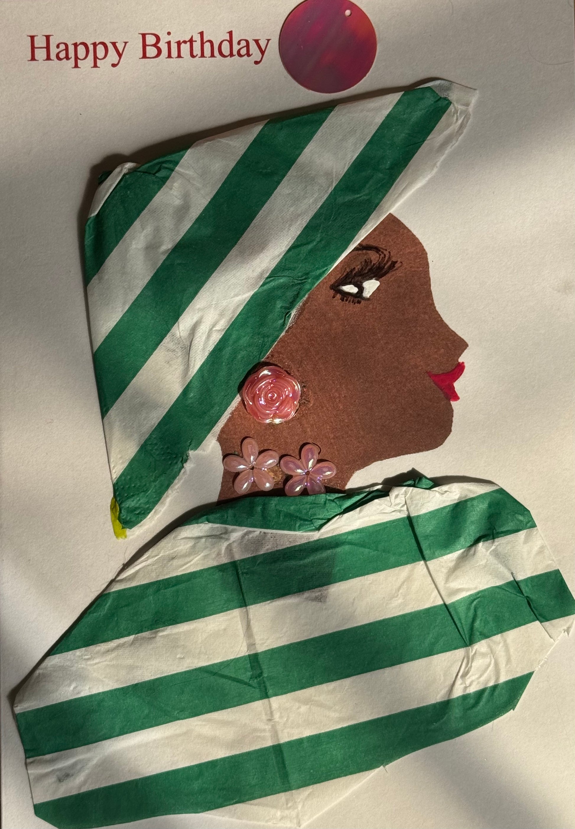 A handmade birthday card showing a woman with dark brown skin in right-facing profile. She wears a green and white striped hat with a matching striped wrap around her shoulders. Her jewelry includes pink flower-shaped earrings and a pink rose stud. A red circular sticker appears above her near the “Happy Birthday” text. The background is white.