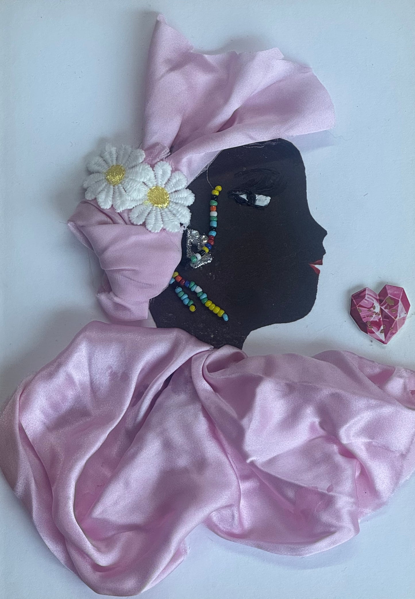 A handmade birthday card showing a side-profile illustration of a woman with dark skin wearing a soft pink satin headwrap and matching satin garment. A white daisy embellishment decorates the side of her head. She wears long multicolored beaded earrings and small gem accents. Her lips are red and her eye is delicately drawn. The background is white with a pink gem sticker near the side.
