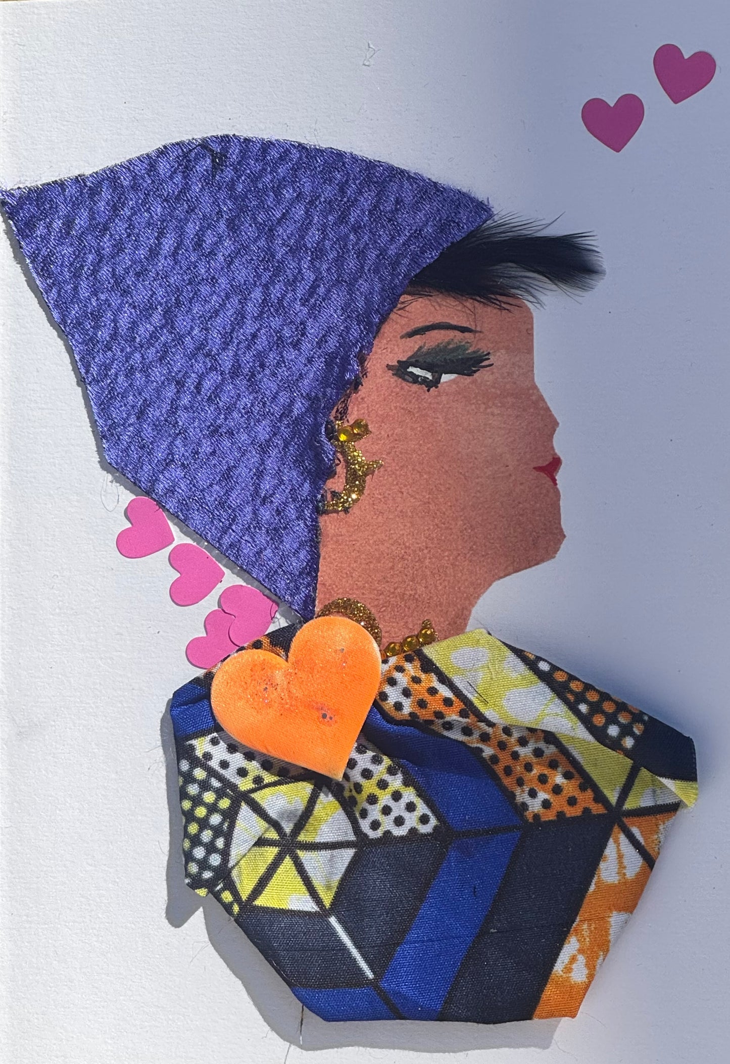 Handmade greeting card that can double as a piece of art featuring a profile of a woman wearing an indigo coloured headwrap in a velvet texture. She also is featured wearing a geometric patterned shawl, with gold detailed jewellery as earrings and a necklace. Above her head to the right of the card, there are two pink decorative hearts placed.