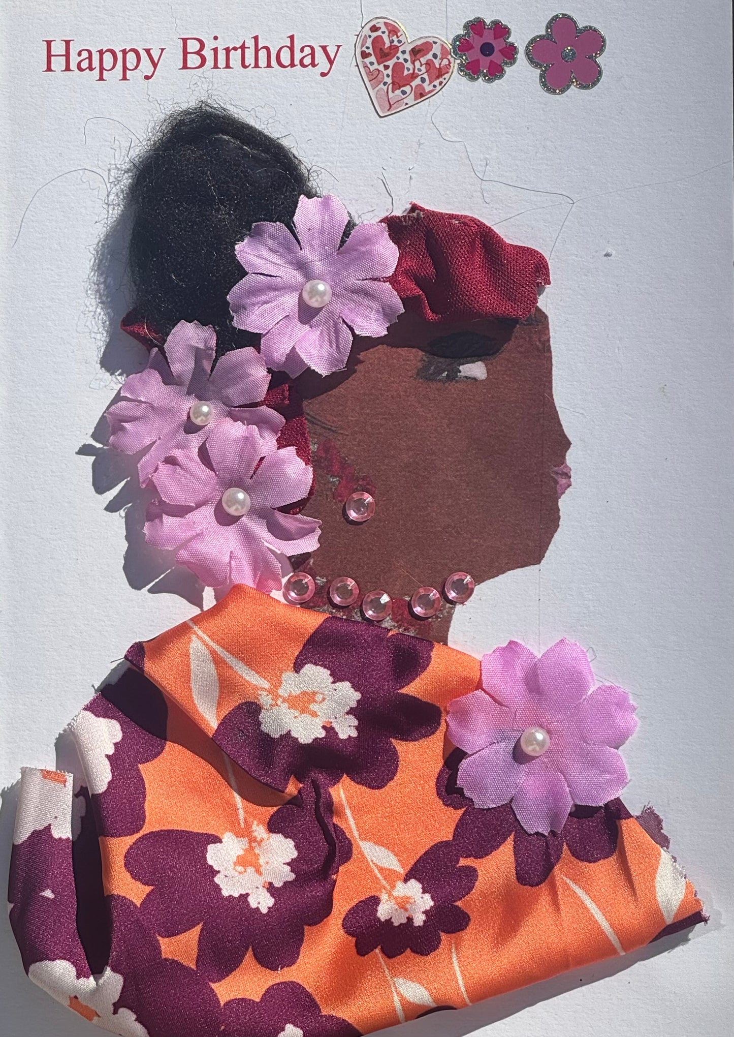 Handmade greeting card that can double as a piece of art featuring a profile of a woman with light purple floral hair accents, pink gem jewelry, and an orange floral patterned garment, with “Happy Birthday” text and decorative pink and purple hearts next to the Happy Birthday and above her head. 