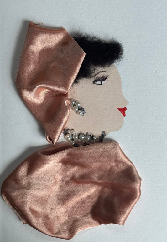 A handmade birthday card showing a woman with light skin in right-facing profile. She wears a blush pink satin headwrap folded into a structured shape. Her jewelry includes pearl drop earrings and a pearl necklace with silver accents. Her shoulders are draped in matching blush satin fabric. She has defined lashes and red lips. “Happy Birthday” is printed in red at the top on a white background.