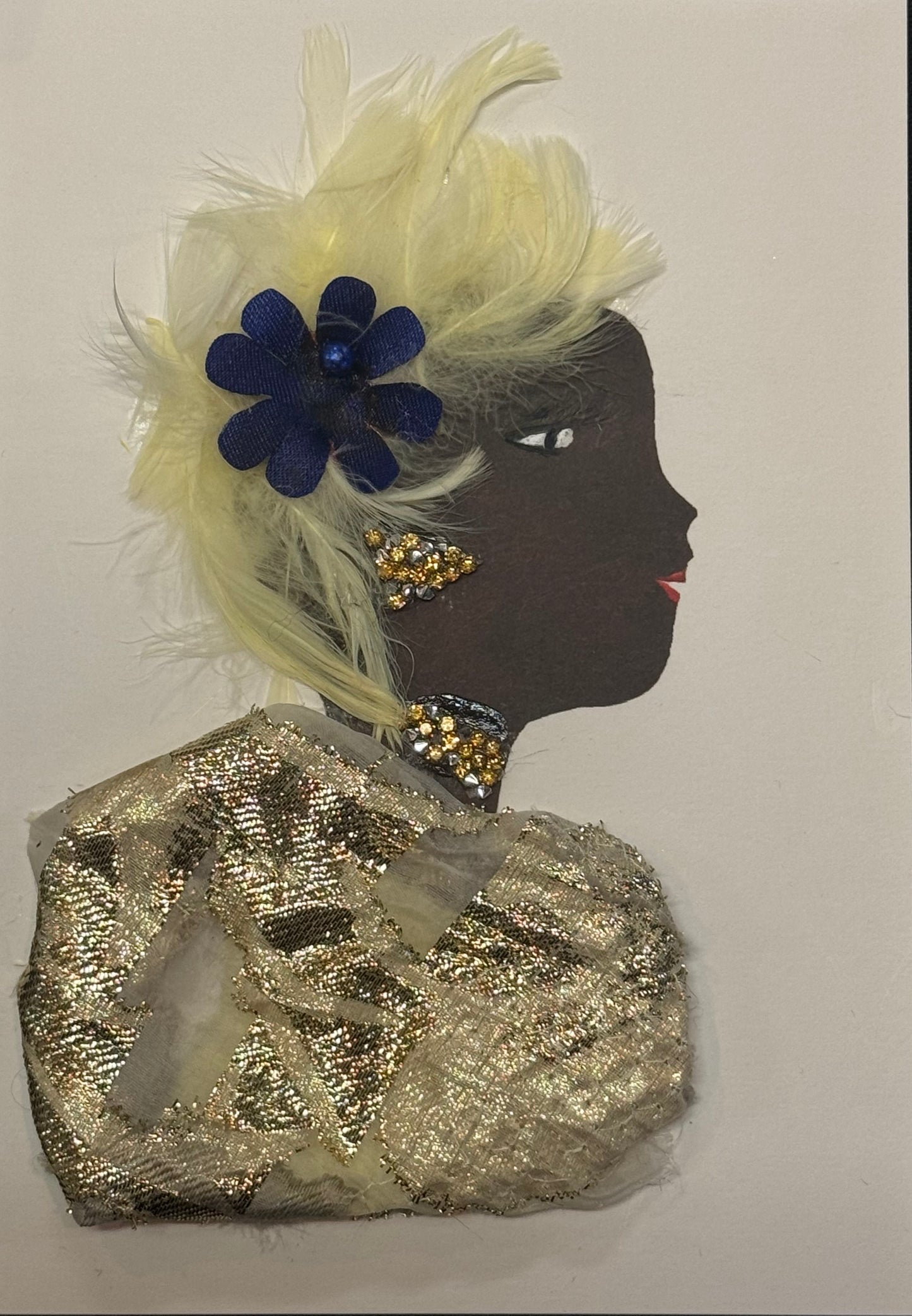 Handmade greeting card that can double as a piece of art featuring a dark brown silhouette of a woman in profile, styled with soft cream-colored feathers forming her hair or headpiece. A deep navy blue flower sits within the feathers, adding contrast. She wears gold-toned textured fabric as clothing and has gold embellishments for earrings and a necklace. Her facial details include a defined eye with lashes and red lips, all layered on a white background with mixed materials including feathers and fabric. 