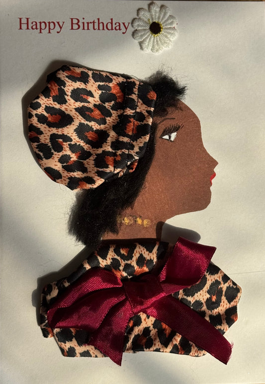 A handmade birthday card showing a woman with dark brown skin in right-facing profile. She wears a leopard print headwrap and a matching leopard print top. A large burgundy satin bow sits across her shoulders. She has subtle gold jewelry and a white flower sticker above her head. “Happy Birthday” appears in red text on a white background.