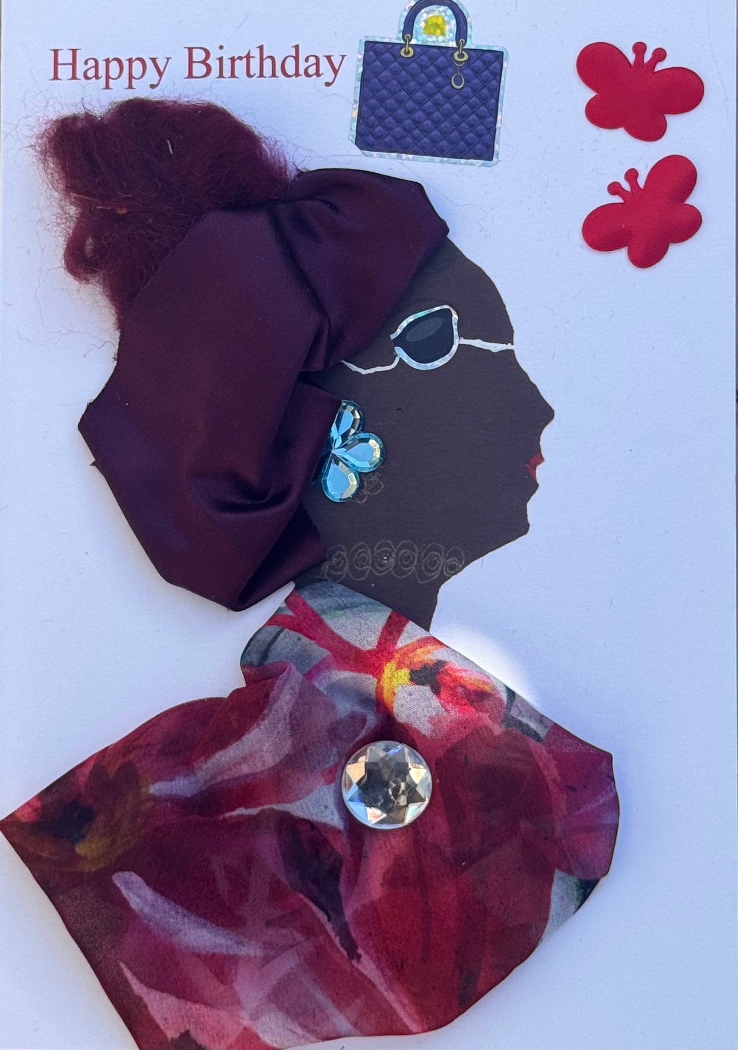 Handmade greeting card that can double as a piece of art featuring a womans profile. She is wearing sunglasses and a burgundy headwrap. She also wears a red, yellow, white, and black floral patterned shawl with a bold silver gem in the centre. For her jewellery accents she wears big silver flower shapped earrings and a gold swirly shaped necklace. Also depicted is a “Happy Birthday” text above her head toward the top left on the card with a purse and two red butterfly stickers next to the text for detail. 
