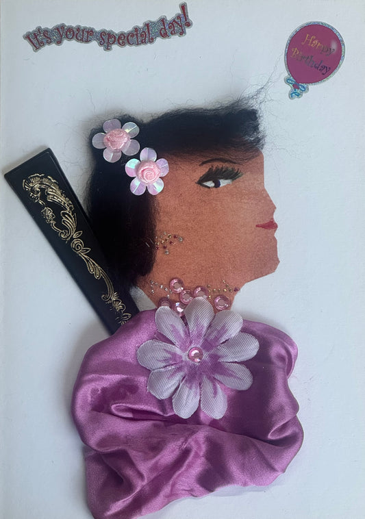 A handmade birthday card featuring a side-profile illustration of a woman wearing a pink satin fabric garment forming her shoulders. Two pink flower embellishments decorate her hair, and she wears rhinestone jewelry around her neck. Her hair is short and dark, with delicate drawn facial features and red lips. The background is white with “It’s your special day!” text above and small decorative stickers.